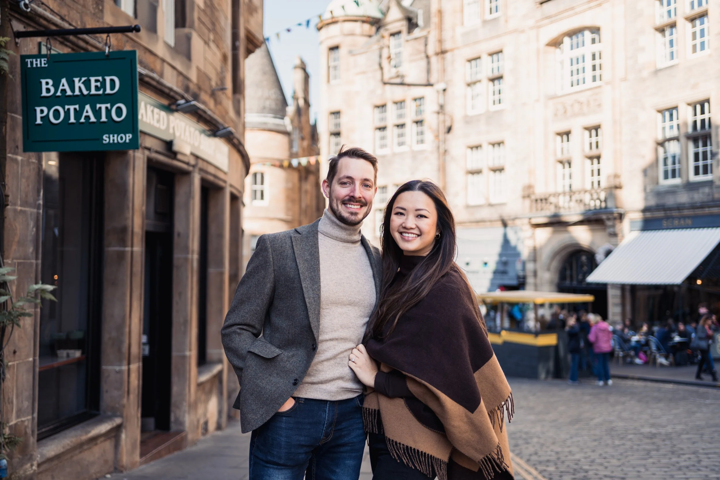 A smiling man and woman stand outdoors on a city street, with historic buildings and a cafe in the background, during daytime.