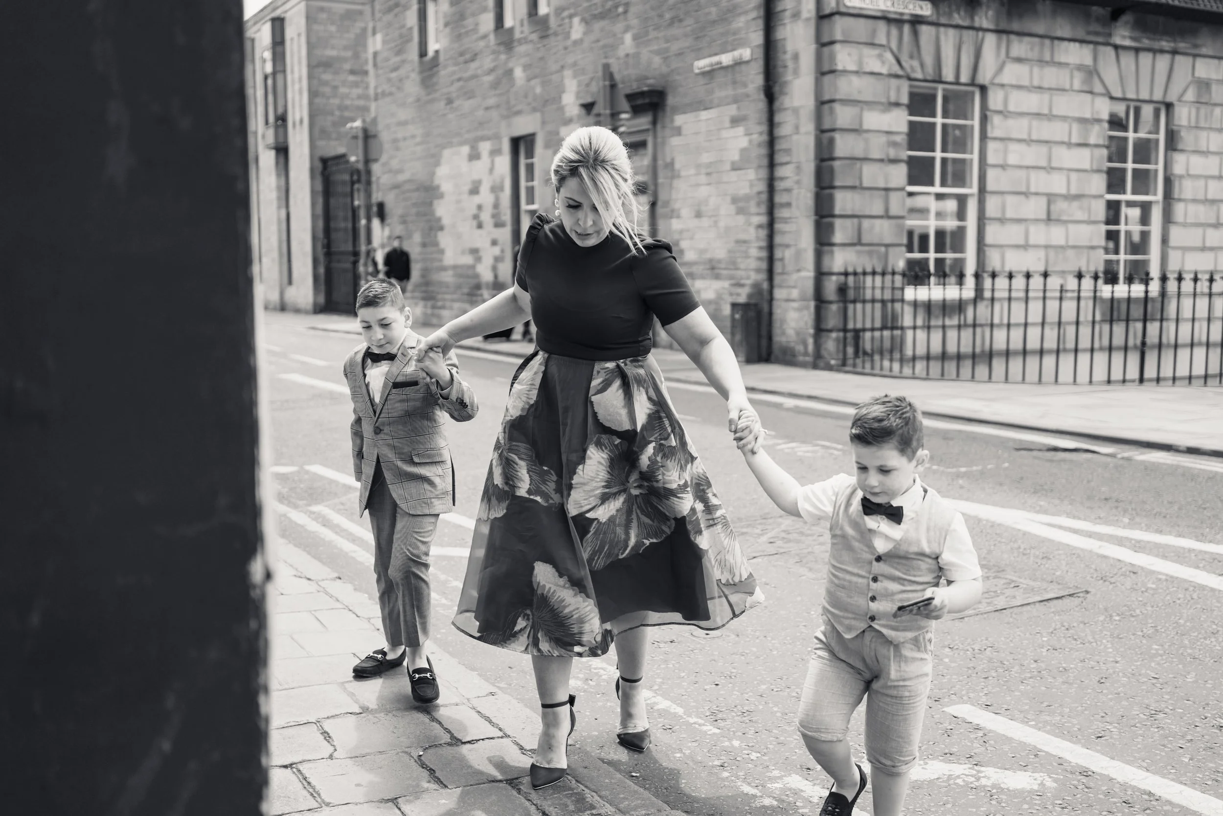 A woman in a floral skirt and high heels walking on a city sidewalk holding the hands of two young boys dressed in formal clothes with bow ties. One boy is holding a phone and the other is adjusting his suit.