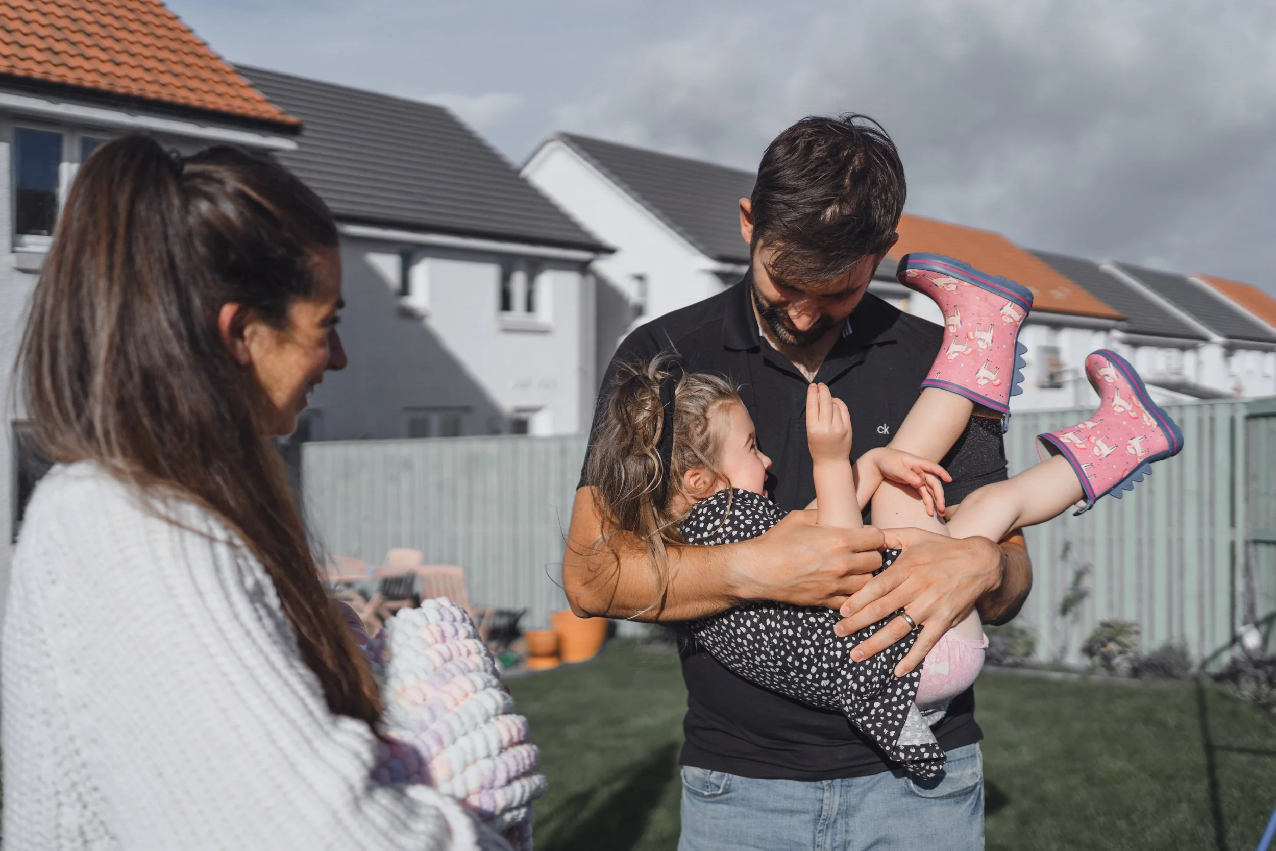 A man holding a young girl in his arms, both smiling, in a backyard with a woman watching and children playing in the background.