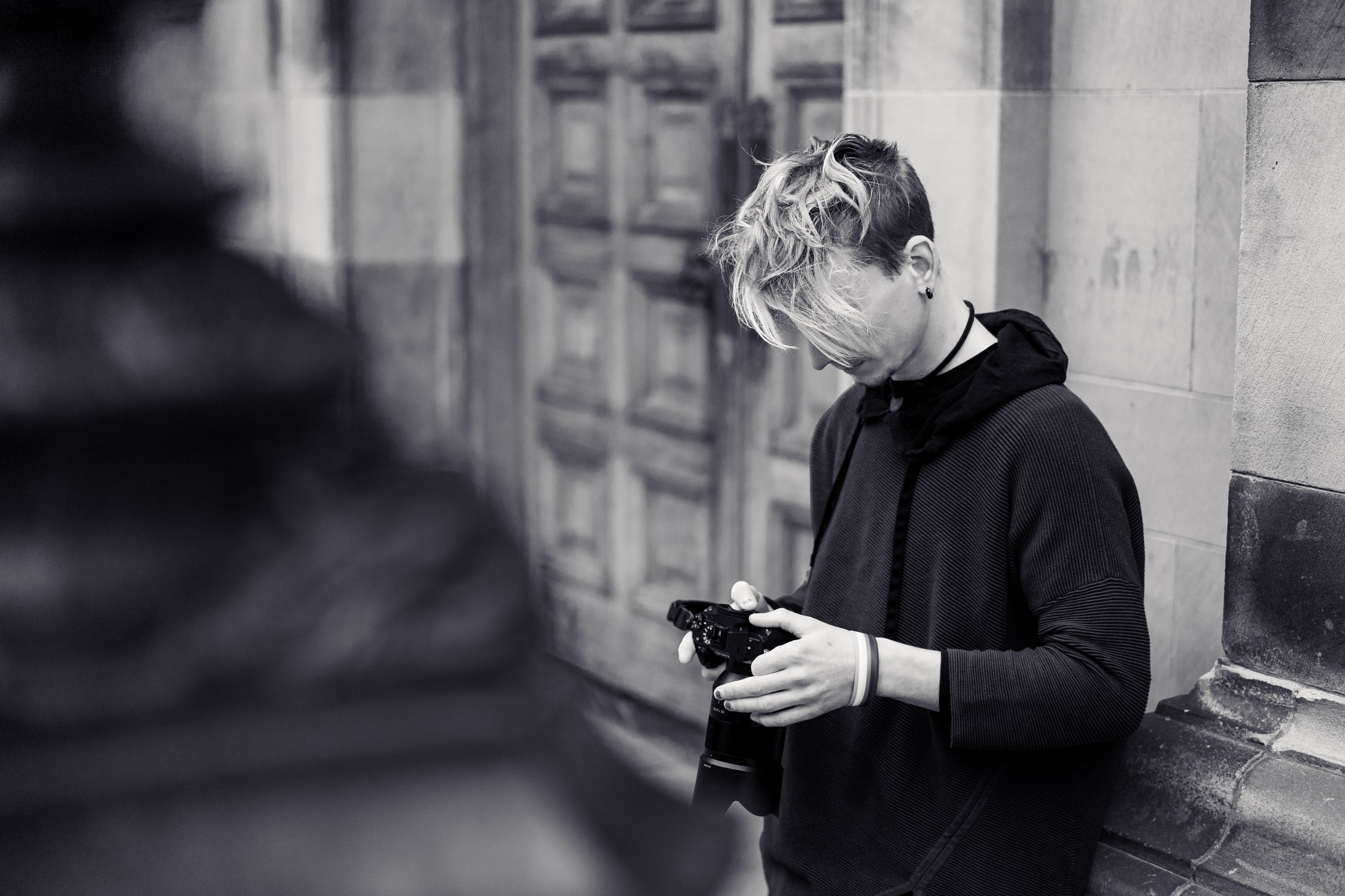 Young man with blonde and dark hair, wearing a dark hoodie, checking a camera while leaning against a stone wall, in an urban setting.