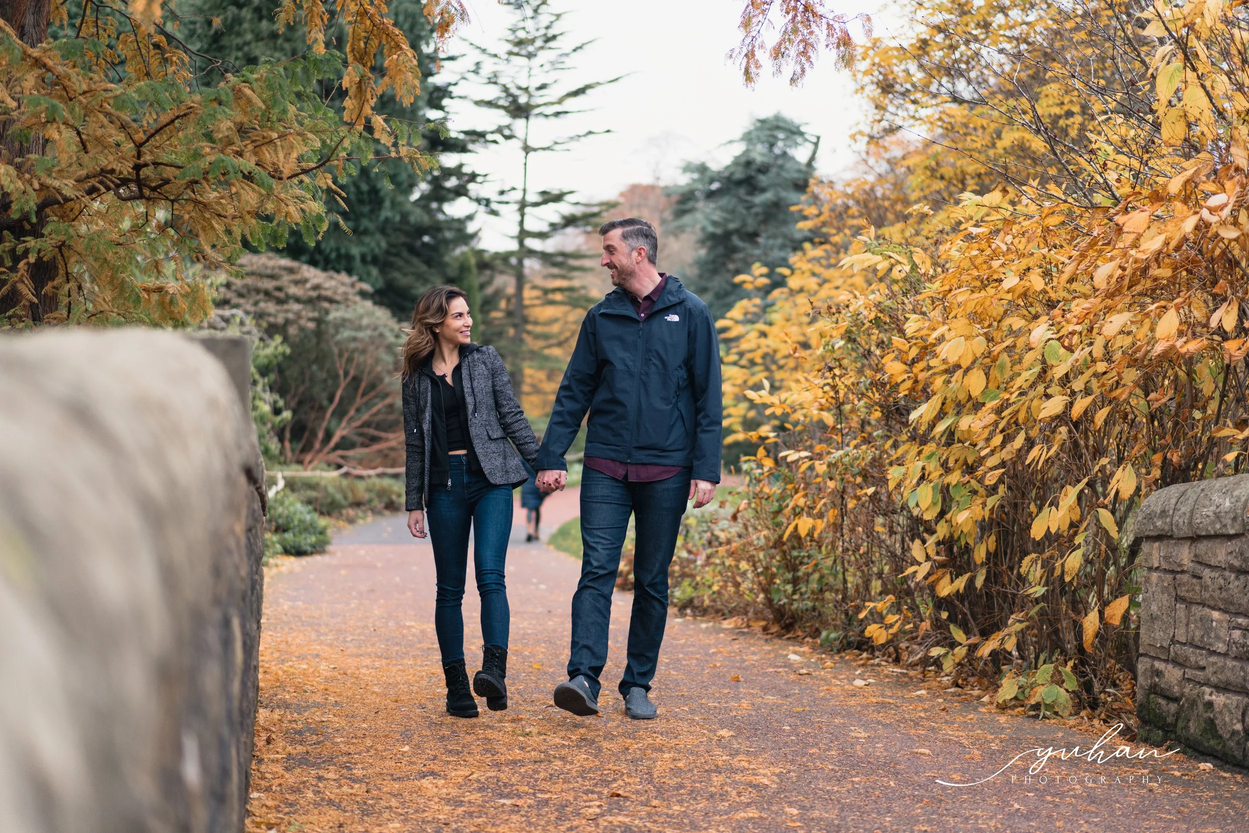 A man and woman walking hand in hand on a fall day, surrounded by yellow and orange leaves, smiling at each other.