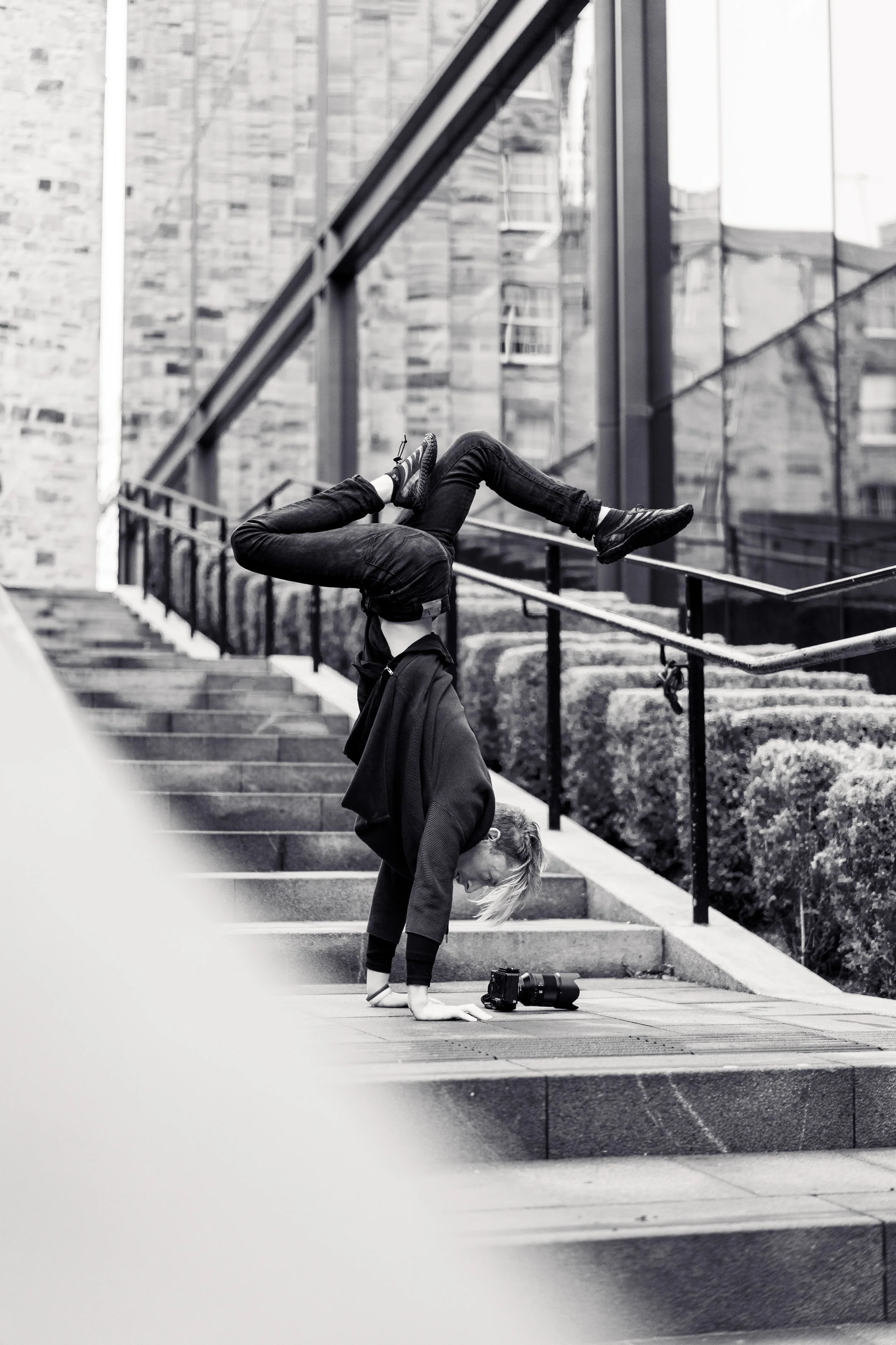 A woman is performing a handstand on outdoor stairs, with her legs bent over her head, in front of a modern glass building. A camera is on the ground nearby.