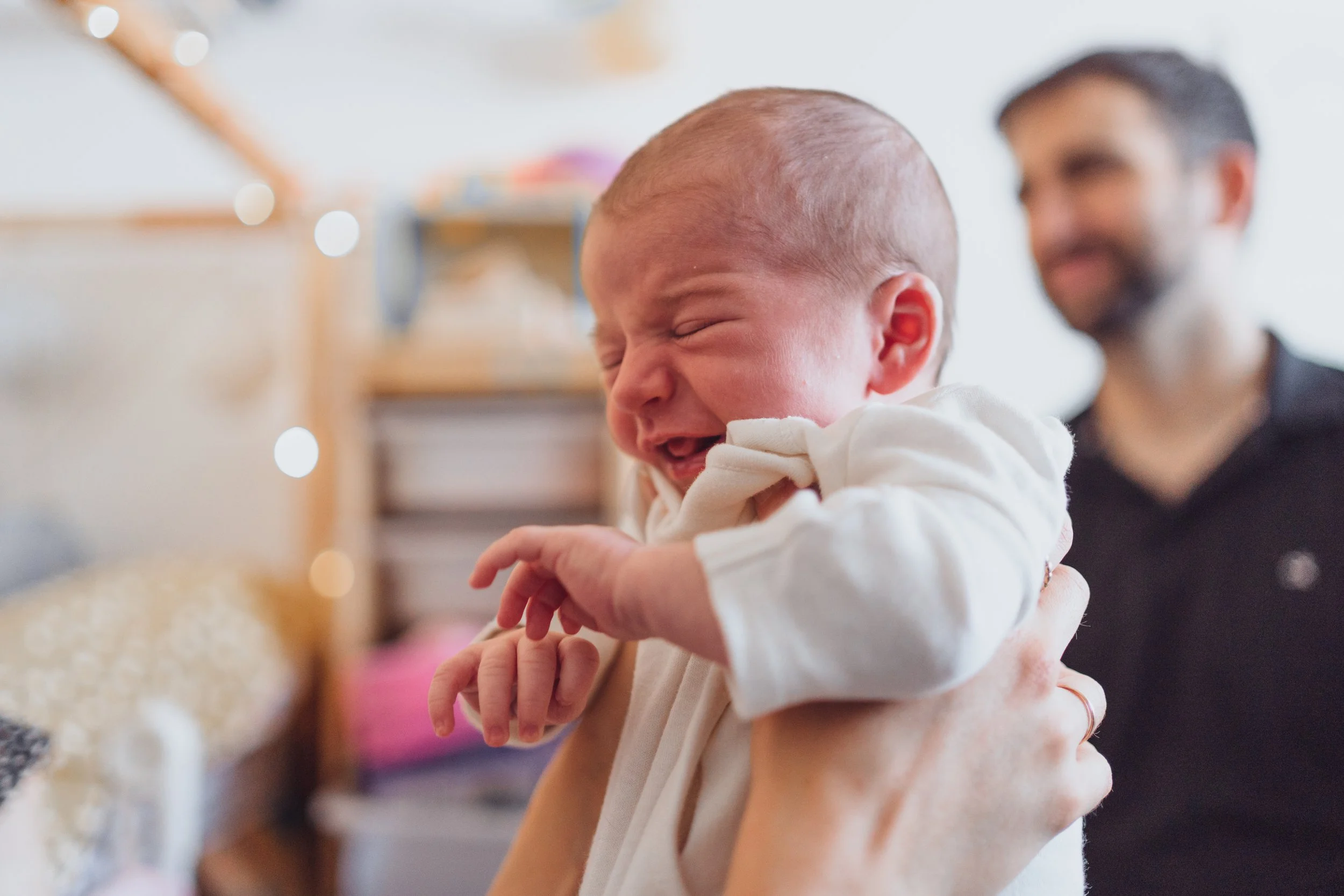A crying baby being held by an adult with an amused man in the background.