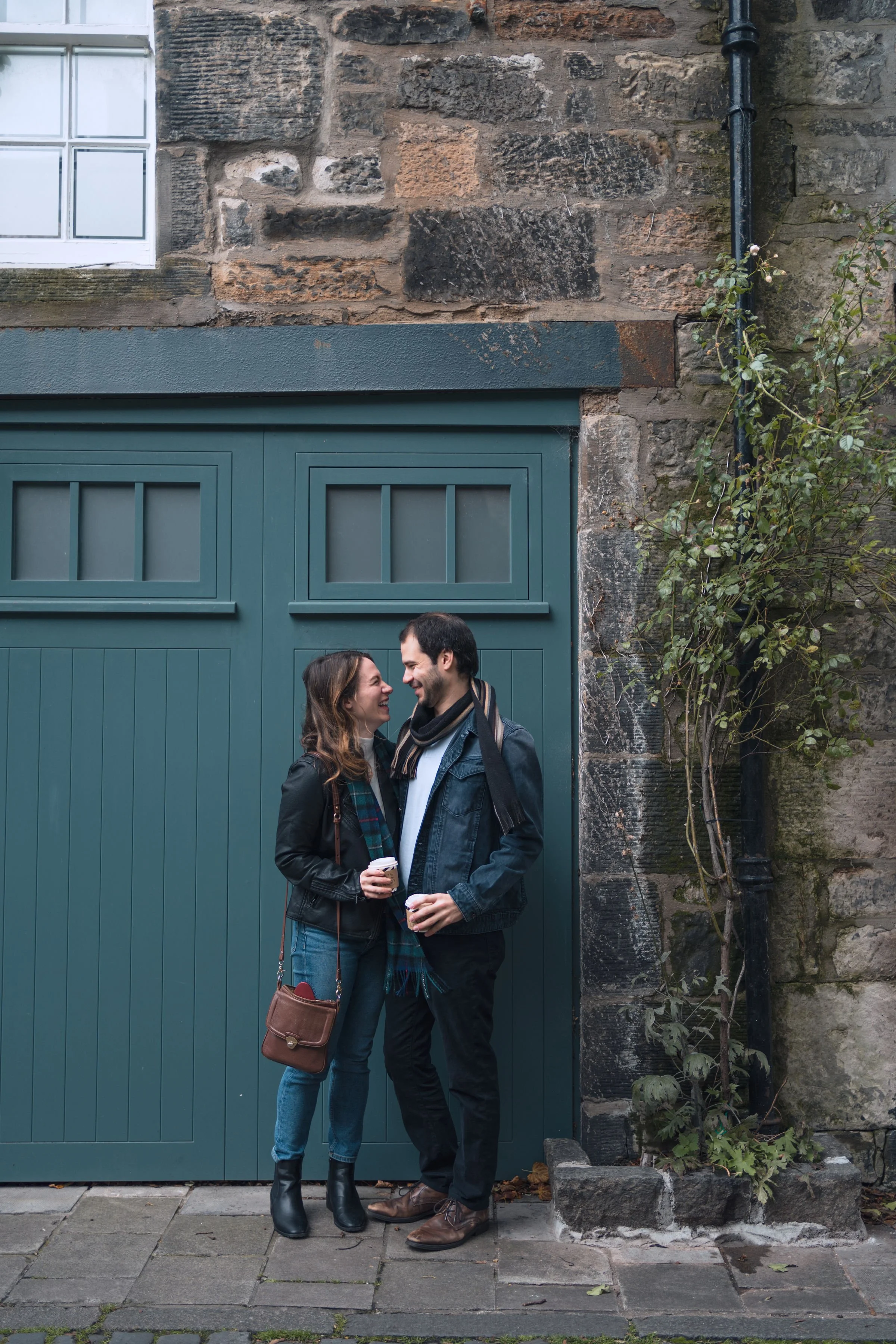 A young couple stands close together, smiling and holding disposable coffee cups, in front of a teal garage door and a stone wall with a window above.