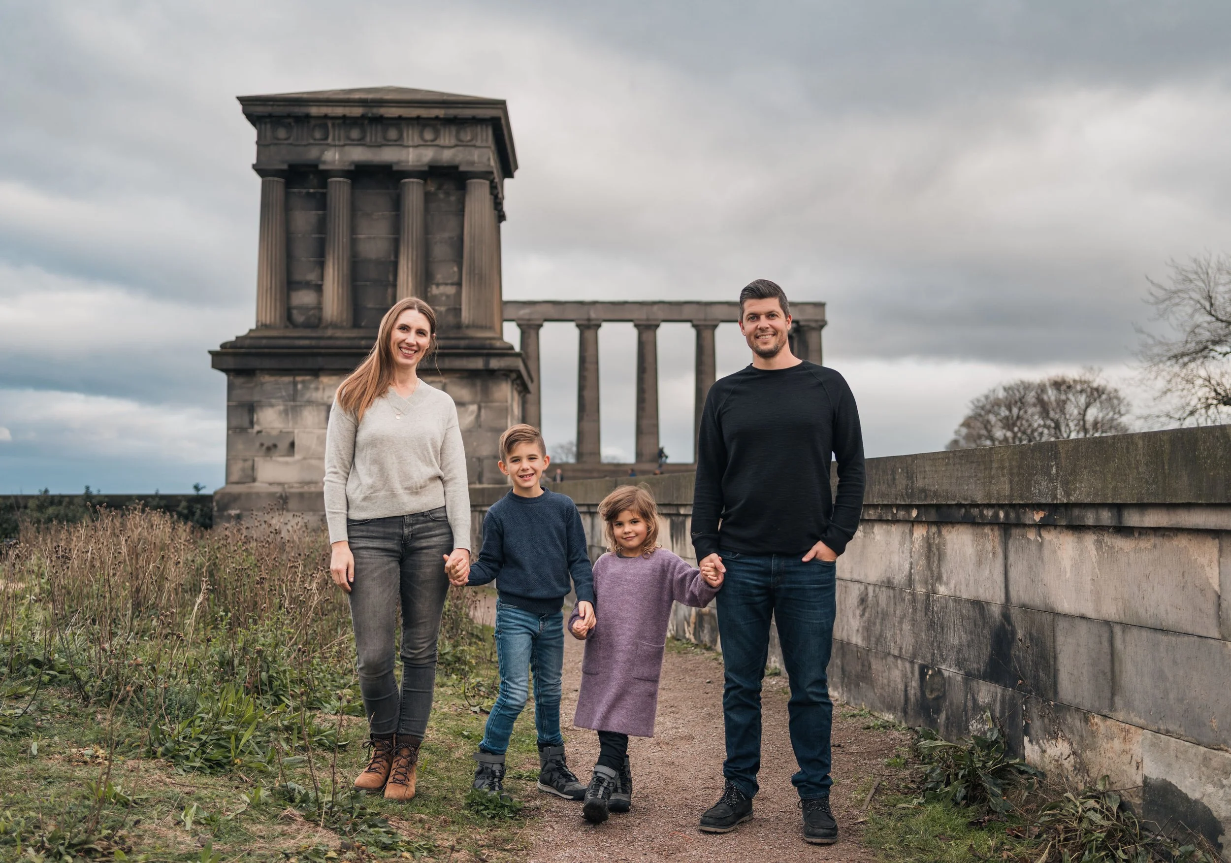 A family of four standing outdoors on a cloudy day in front of a historic monument with columns, holding hands and smiling.