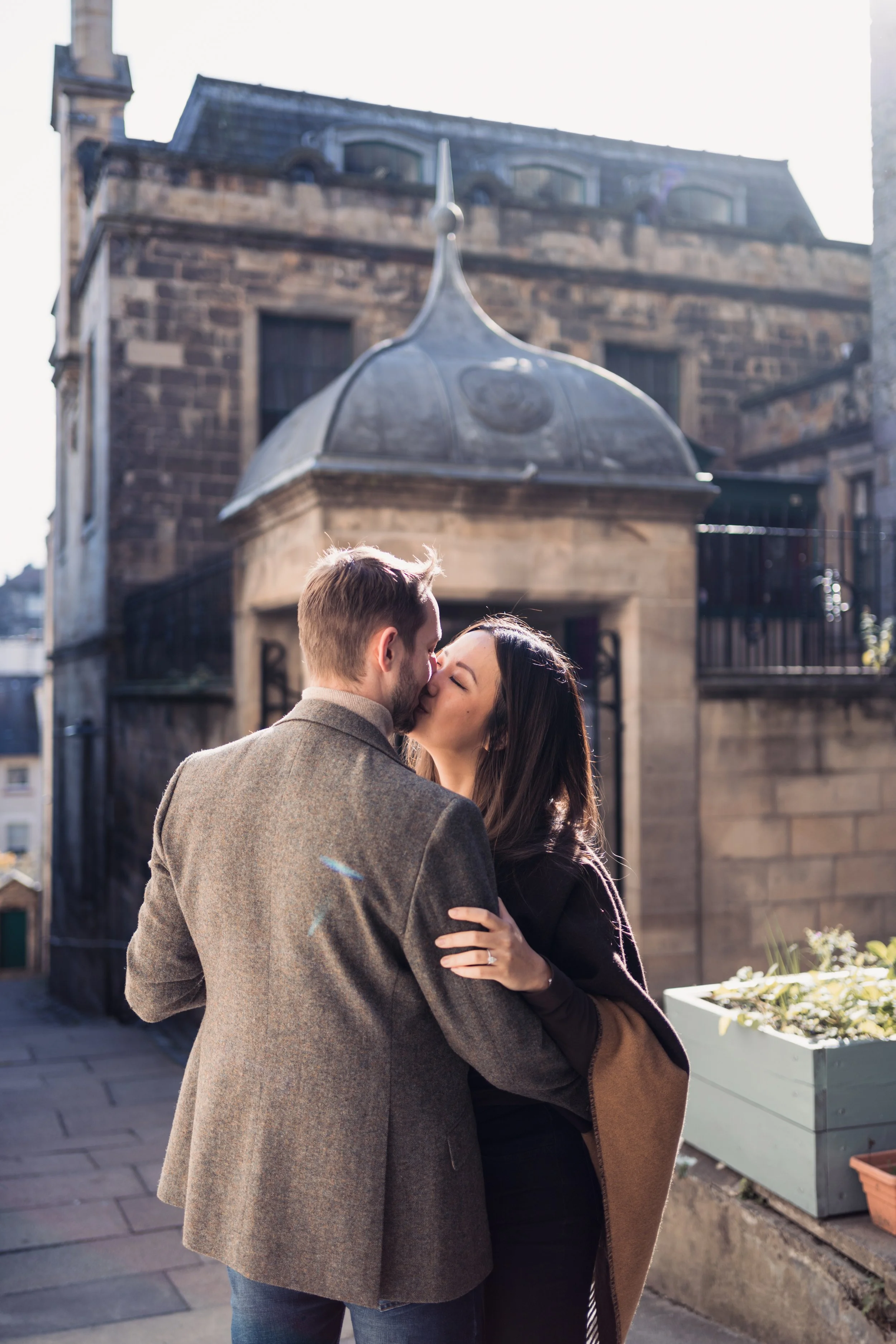 A couple sharing a kiss outdoors, with a historic building in the background.