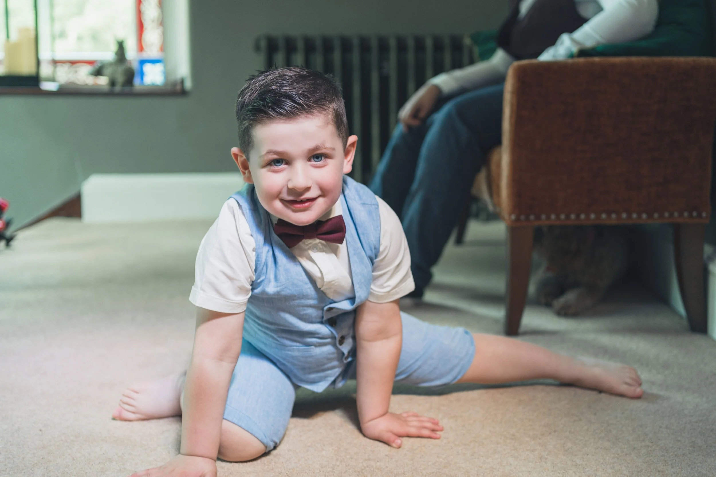 A young boy with dark hair, wearing a white shirt, a bow tie, and a blue vest and shorts, is performing a split on a carpeted floor, smiling at the camera.
