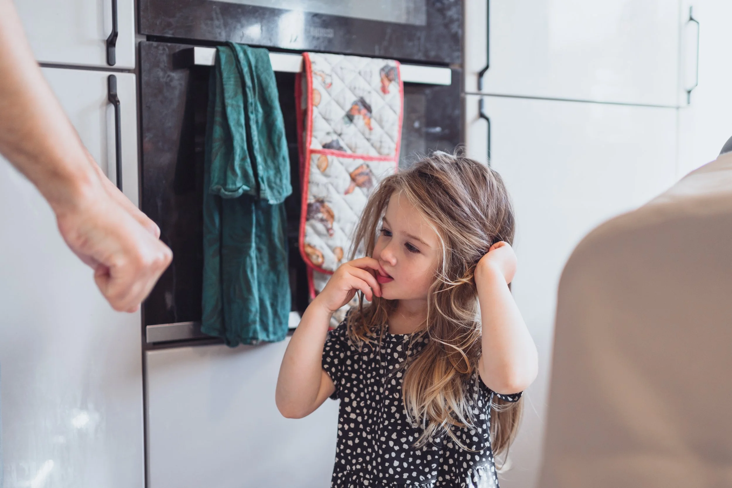 A young girl with long light brown hair, wearing a black dress with white polka dots, appears concerned or distracted as she stands in a kitchen. In the background, there are kitchen cabinets, and hanging on a rack are two dish towels, one green and 