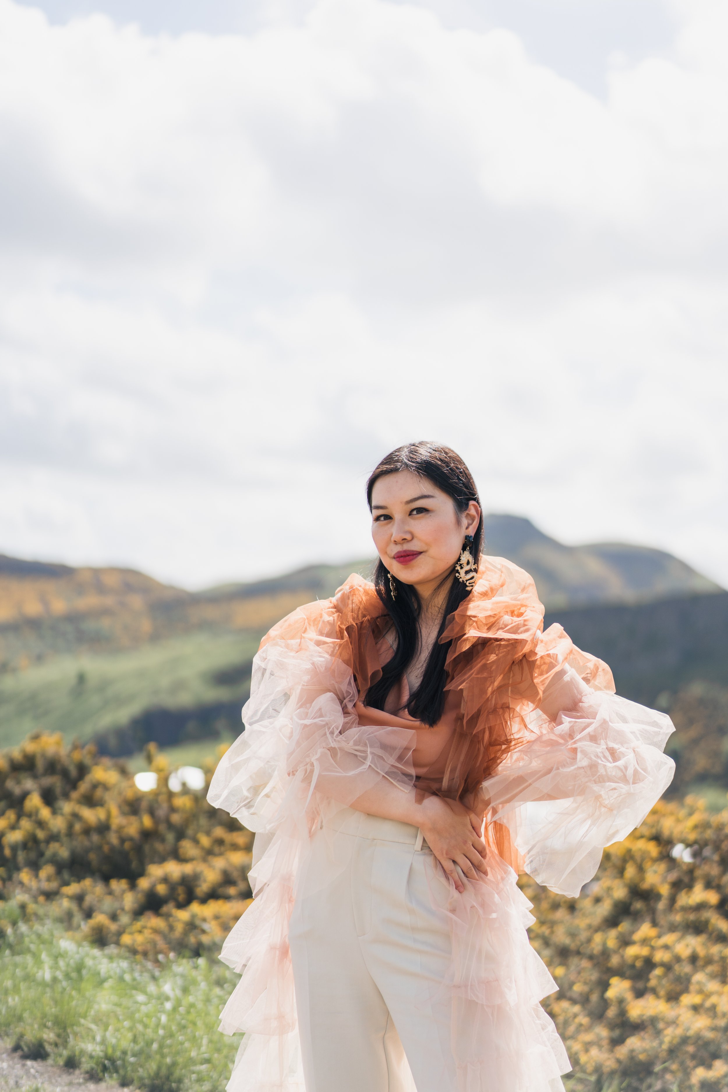 A woman standing outdoors in front of a scenic landscape with hills and trees, wearing a light-colored outfit with ruffled, sheer, orange and white fabric, and large earrings.