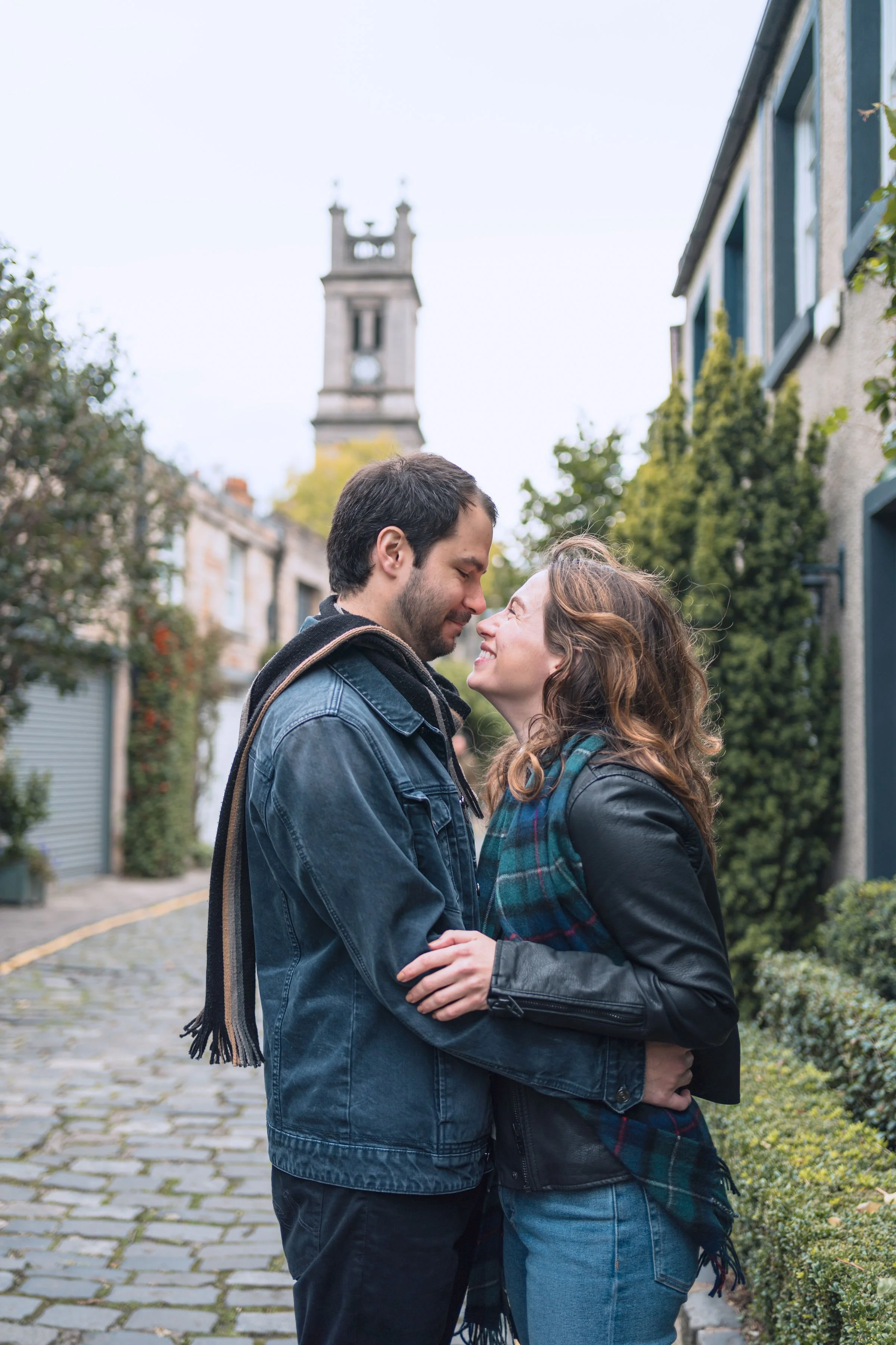 A young couple embraces on a cobblestone street with buildings and a clock tower in the background.