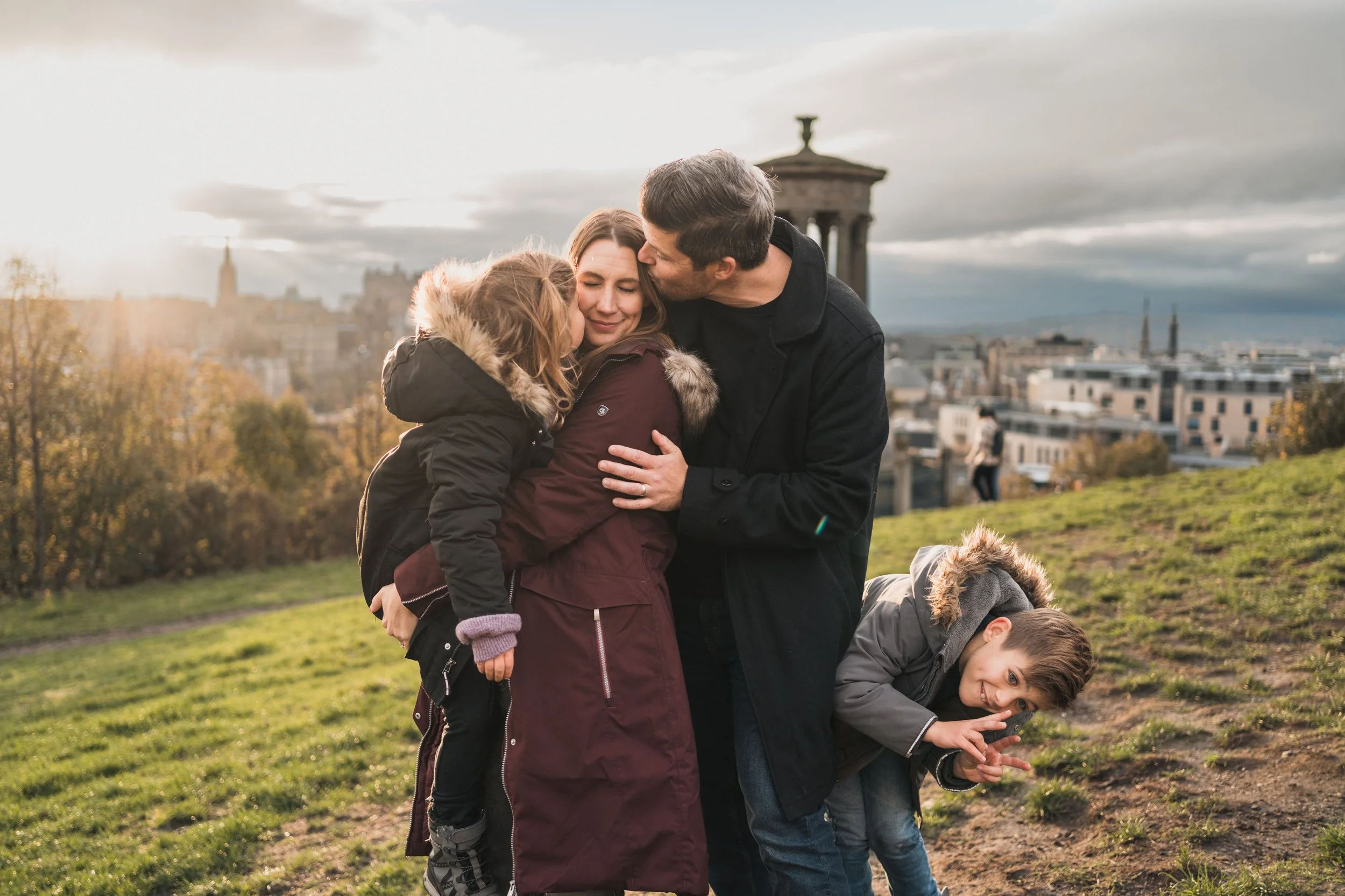 A family of four, including a man, woman, girl, and boy, embracing and smiling on a grassy hill with a cityscape in the background during sunset.