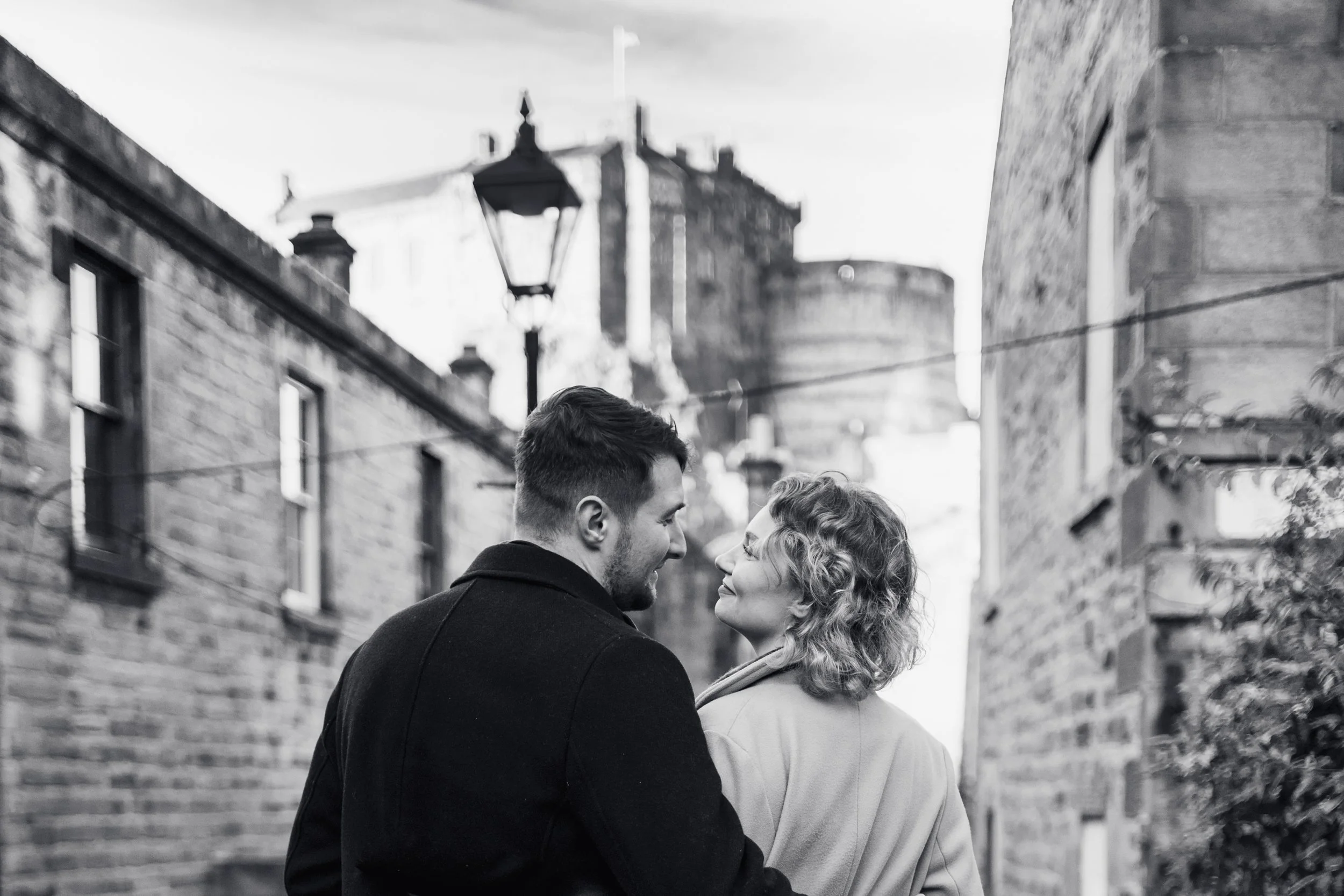A young man and woman stand closely together on a city street, looking into each other's eyes and smiling, with old brick buildings, a street lamp, and a castle in the background.