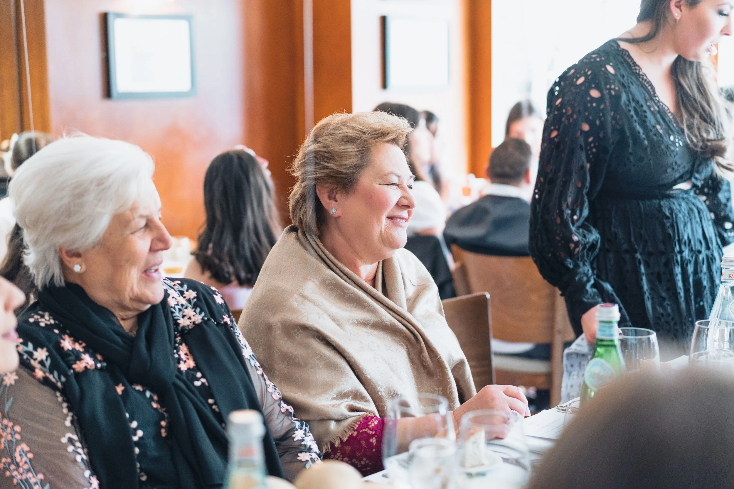 Women sitting at a dining table during a social event, with one woman smiling and another woman on the right standing, wearing a black lace dress, in a room with wood-paneled walls.
