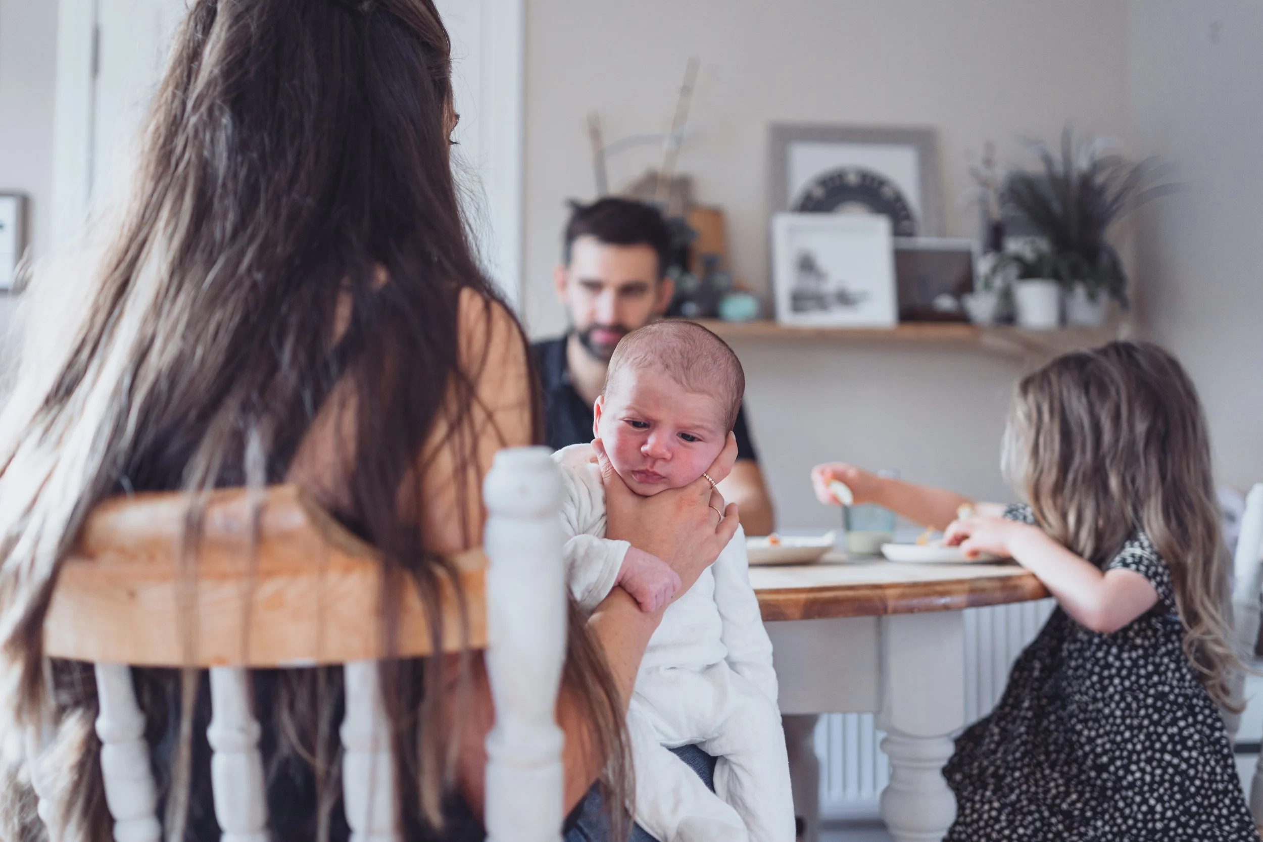 A woman holding a baby with a concerned expression, a man sitting behind them, and a young girl eating at a dining table in a cozy home setting.