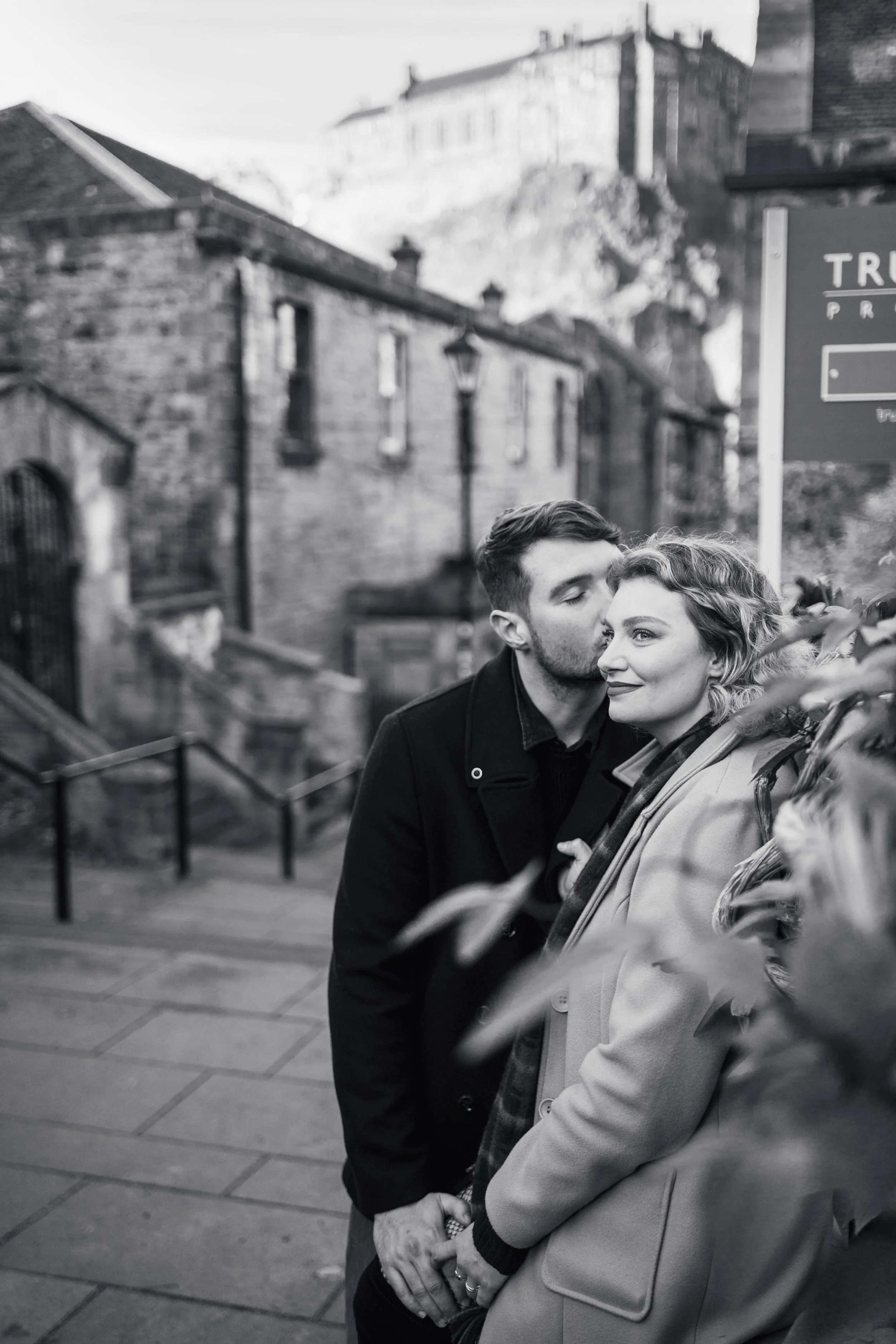 A black-and-white photo of a couple standing close together on a city street with old brick buildings and stairs in the background; the man is kissing the woman's temple as she looks ahead with a smile.