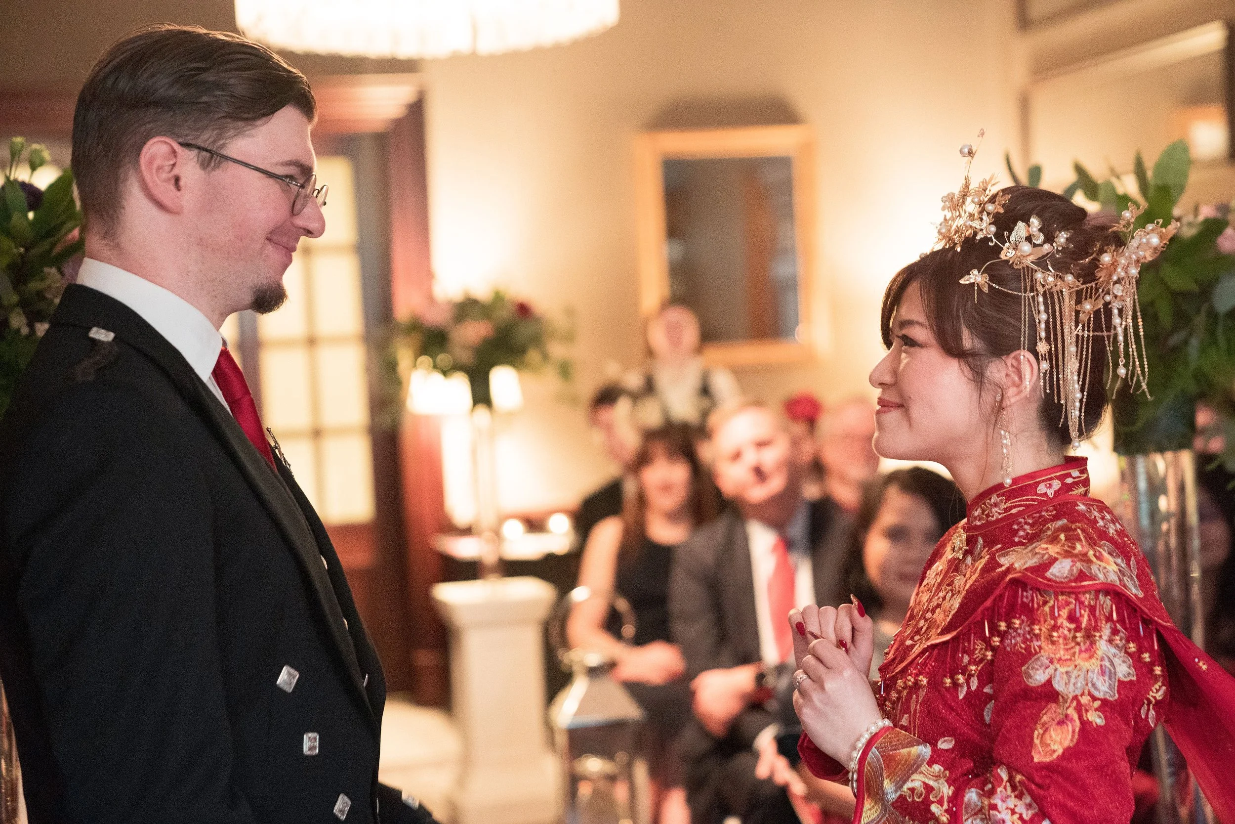 A couple getting married, the groom in a military uniform and the bride in a traditional red Chinese wedding dress with intricate golden embroidery and ornate hair accessories, smiling at each other during a wedding ceremony with guests in the backgr