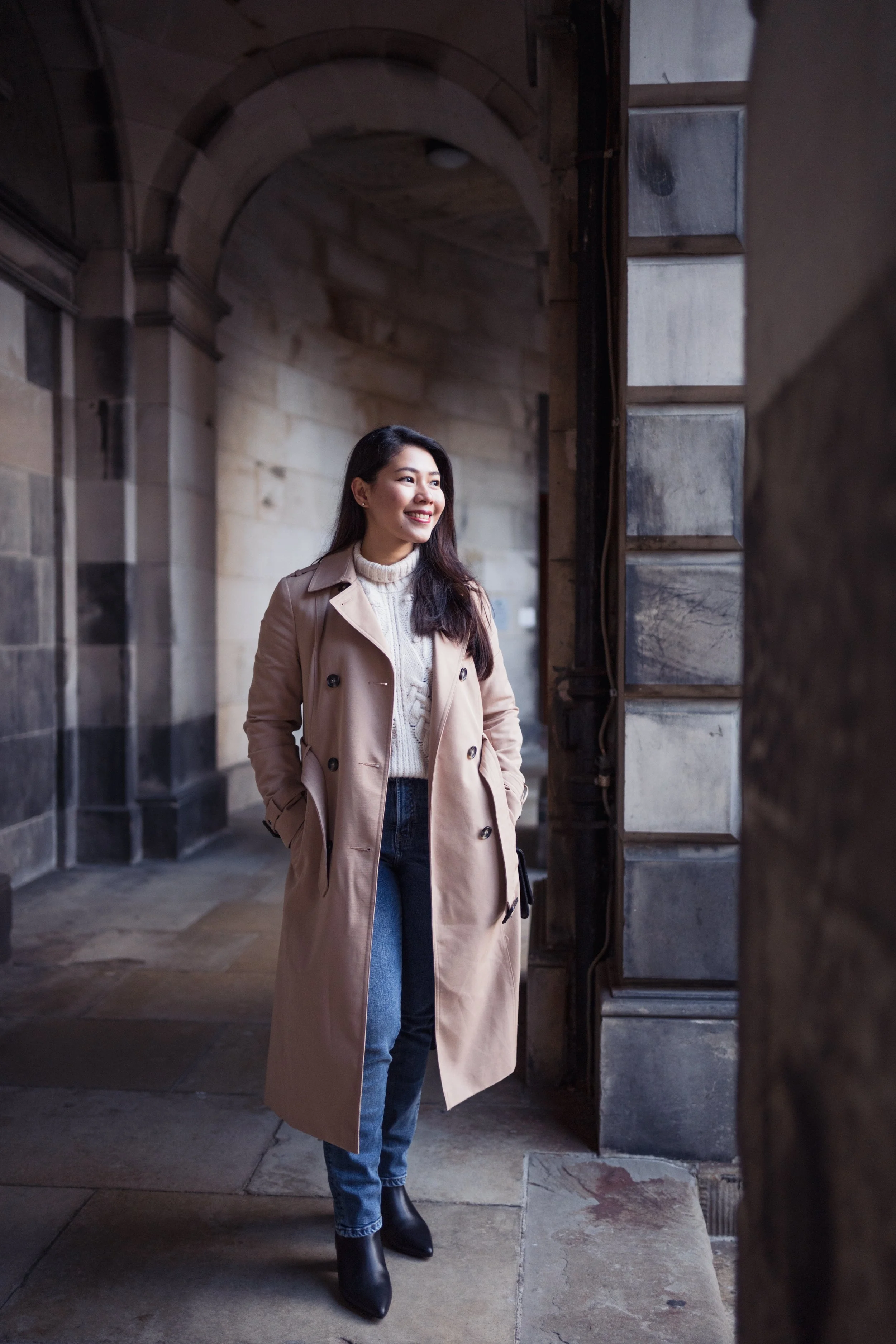 A woman in a beige trench coat, white sweater, blue jeans, and black boots standing in an arched stone passageway, smiling and looking to the side.