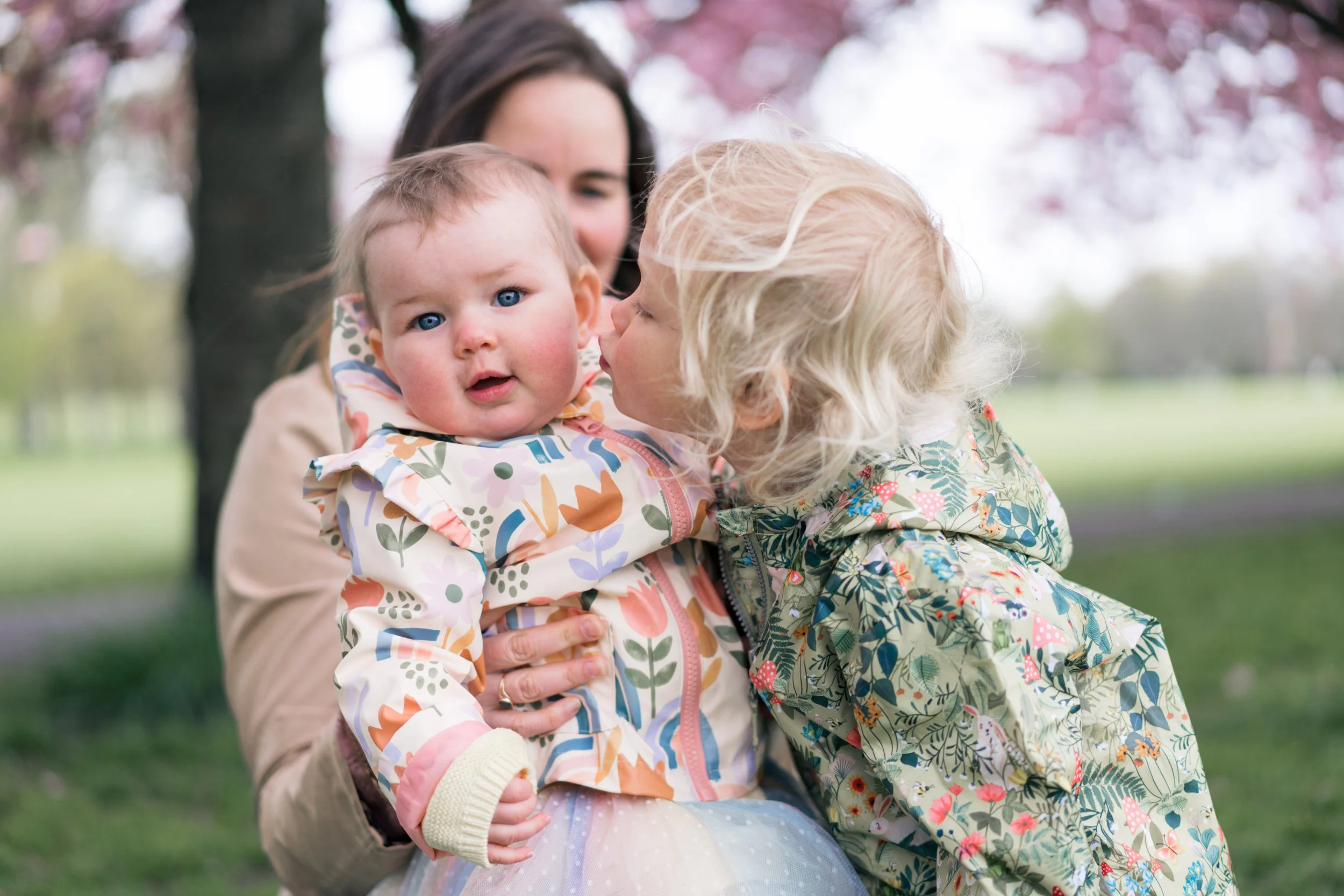 A woman holding a baby in her arms while a young girl leans in to kiss the baby on the cheek outdoors with a background of pink blossoming trees.