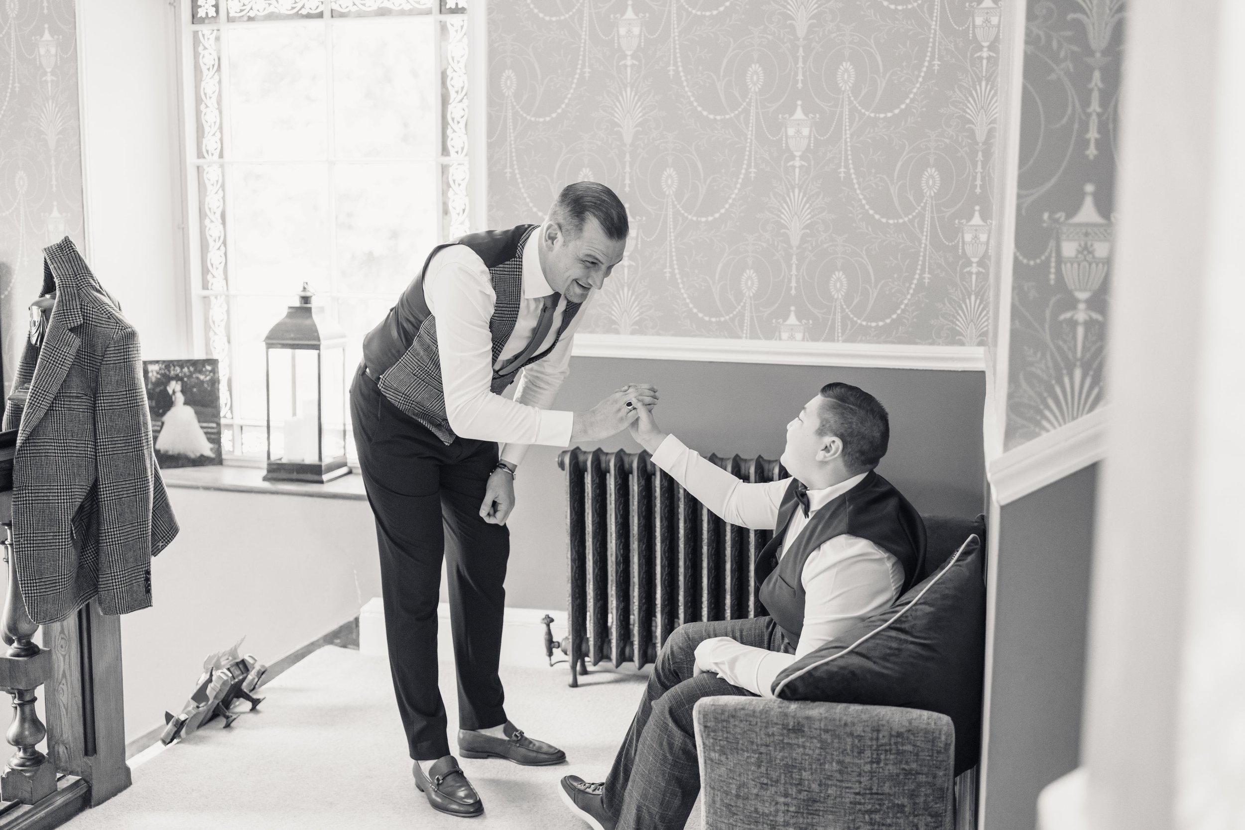Two men in formal attire sharing a handshake and smile in a bright room, one sitting on a sofa and the other standing, with decor and a window in the background.
