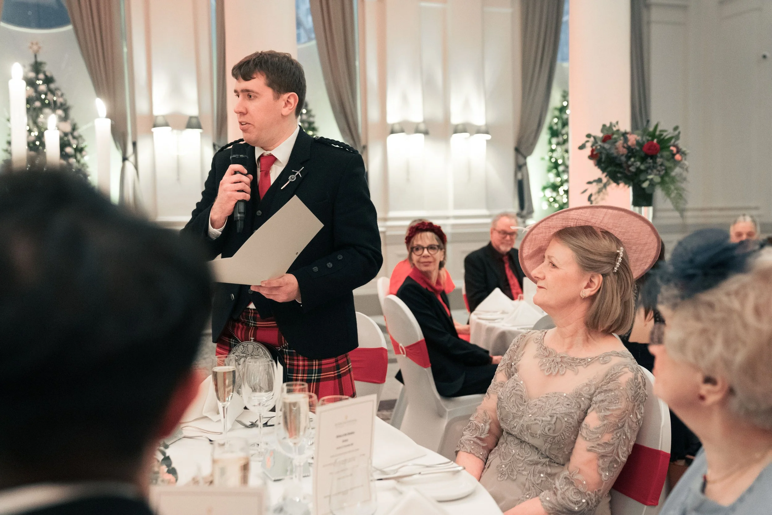 A man in formal attire giving a speech at a wedding reception, surrounded by elegantly dressed guests in a decorated banquet hall.