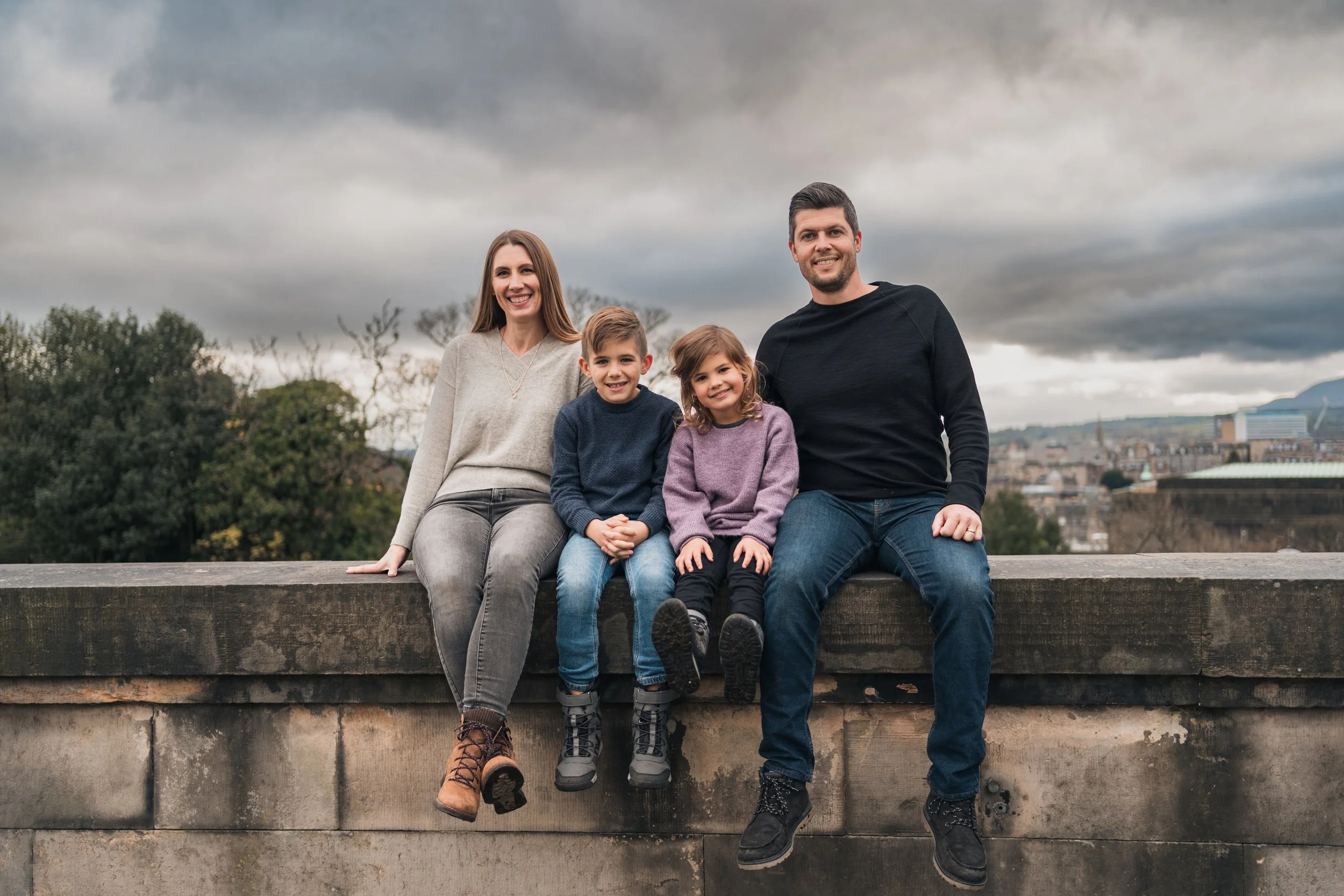 Family of four sitting on a stone wall outdoors, smiling, with dark cloudy sky and cityscape in the background.