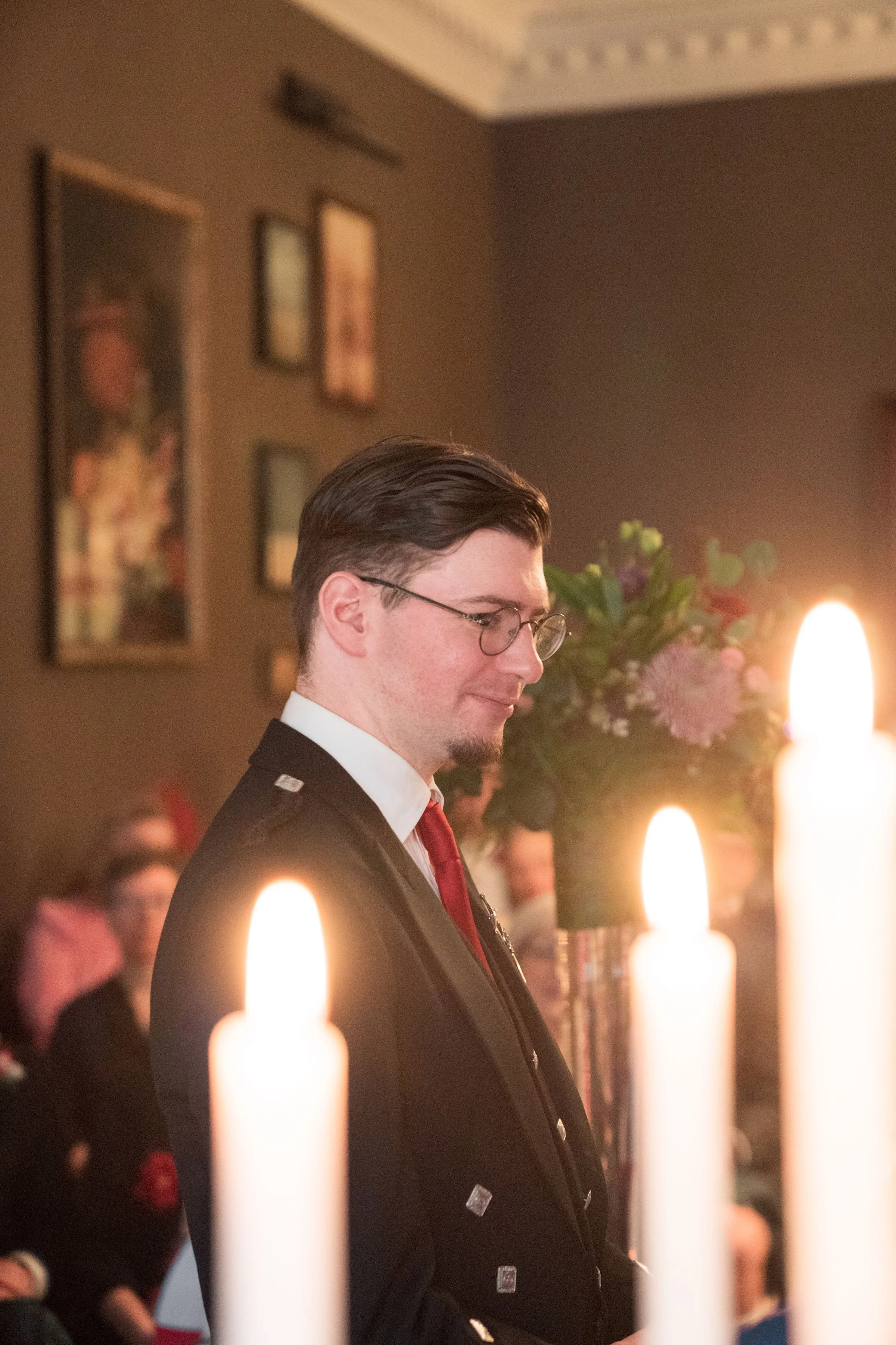 A man with glasses and dark hair, wearing a formal suit and red tie, standing indoors near lit candles and a flower arrangement, with framed pictures on the wall behind him.
