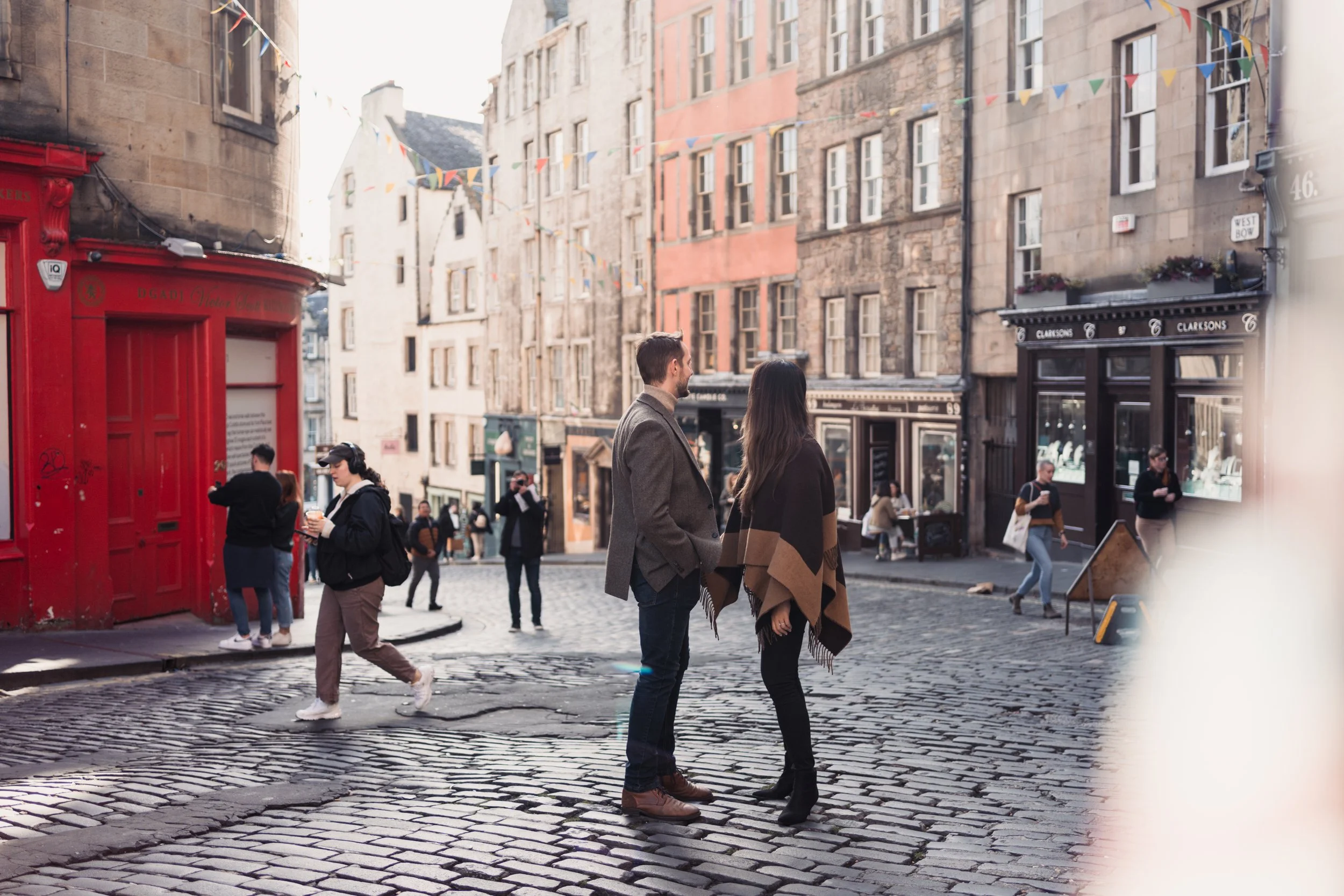 A man and a woman having a conversation on a cobblestone street in a city, with several people walking by and storefronts in the background.