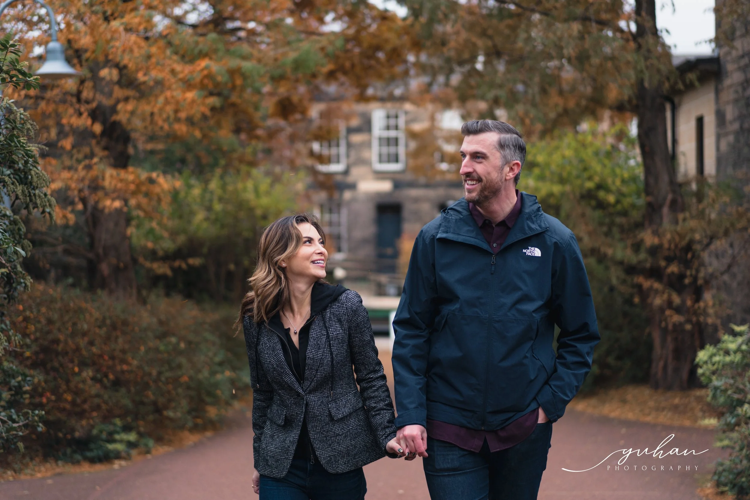 A smiling couple walking hand in hand on a path surrounded by trees with autumn-colored leaves, in a park or garden setting.