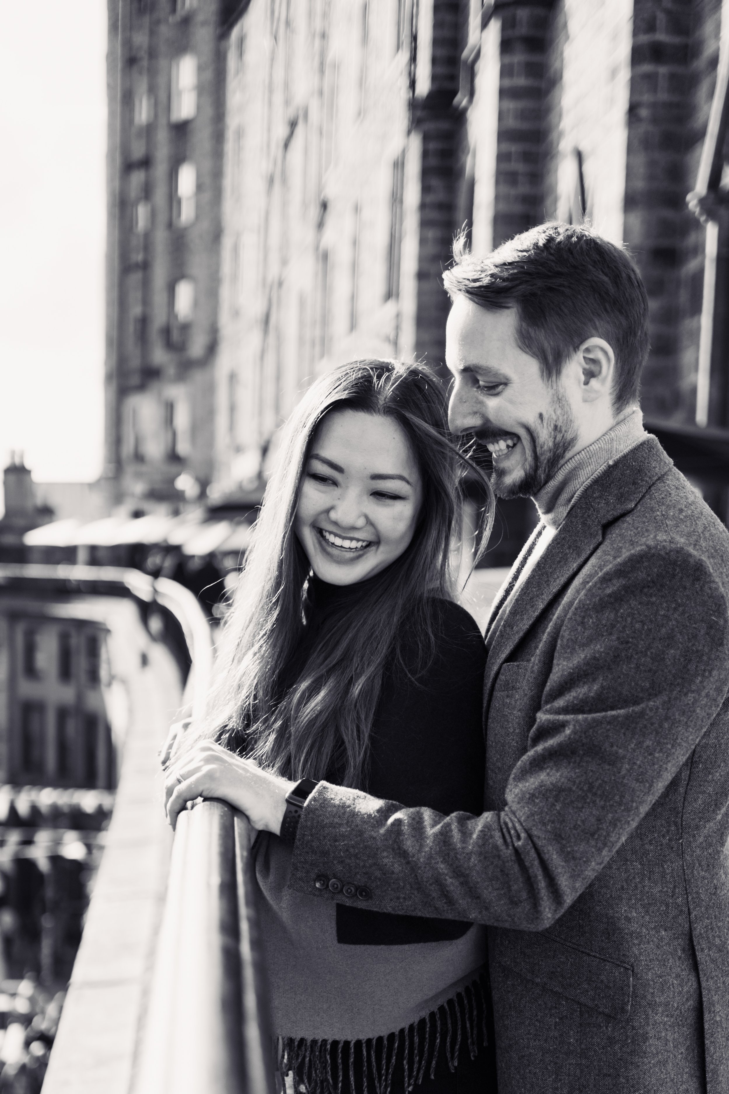 A smiling man and woman leaning on a railing outdoors in a city setting, with buildings in the background.