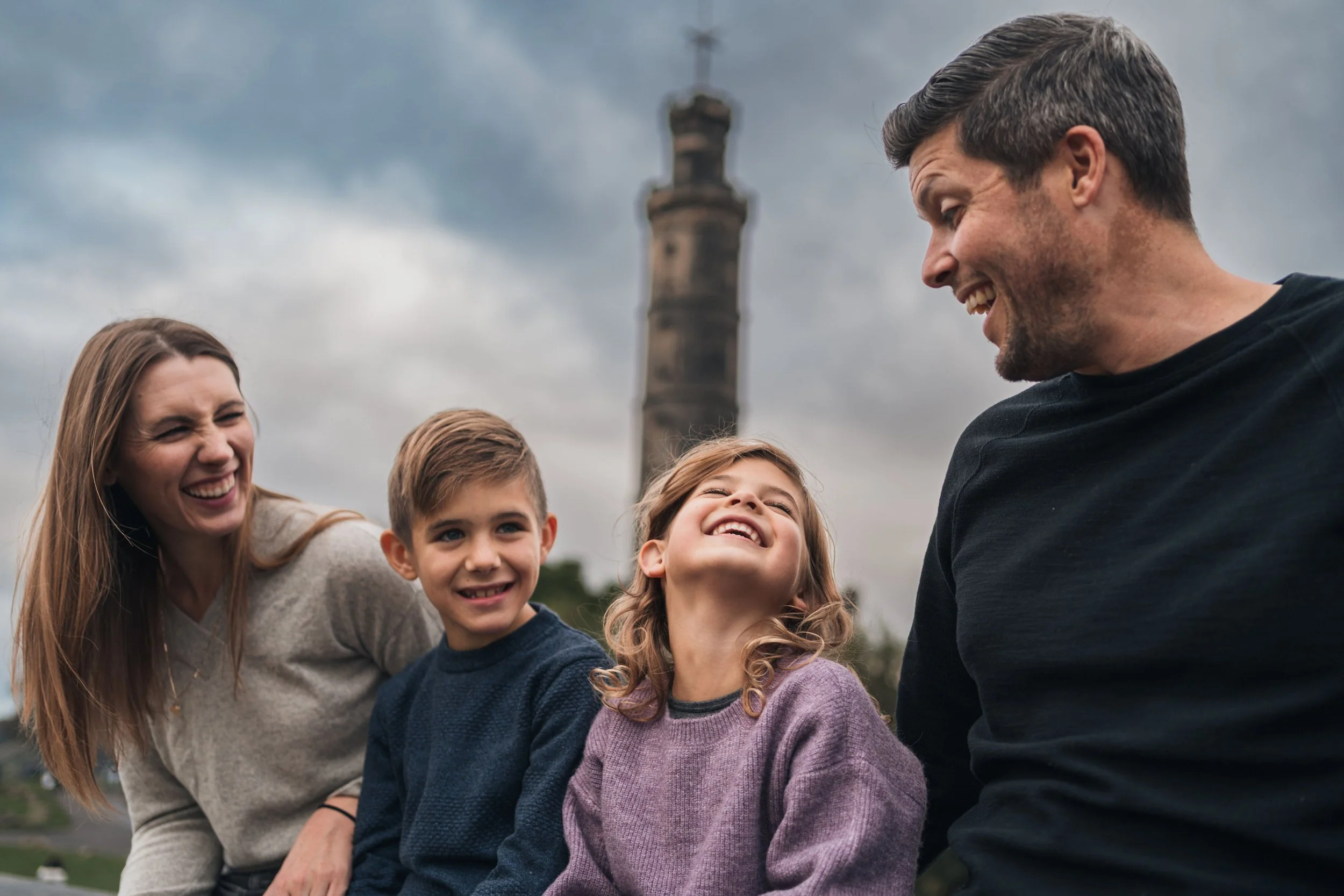 Family of four smiling and laughing outdoors with a tall monument in the background under cloudy sky.
