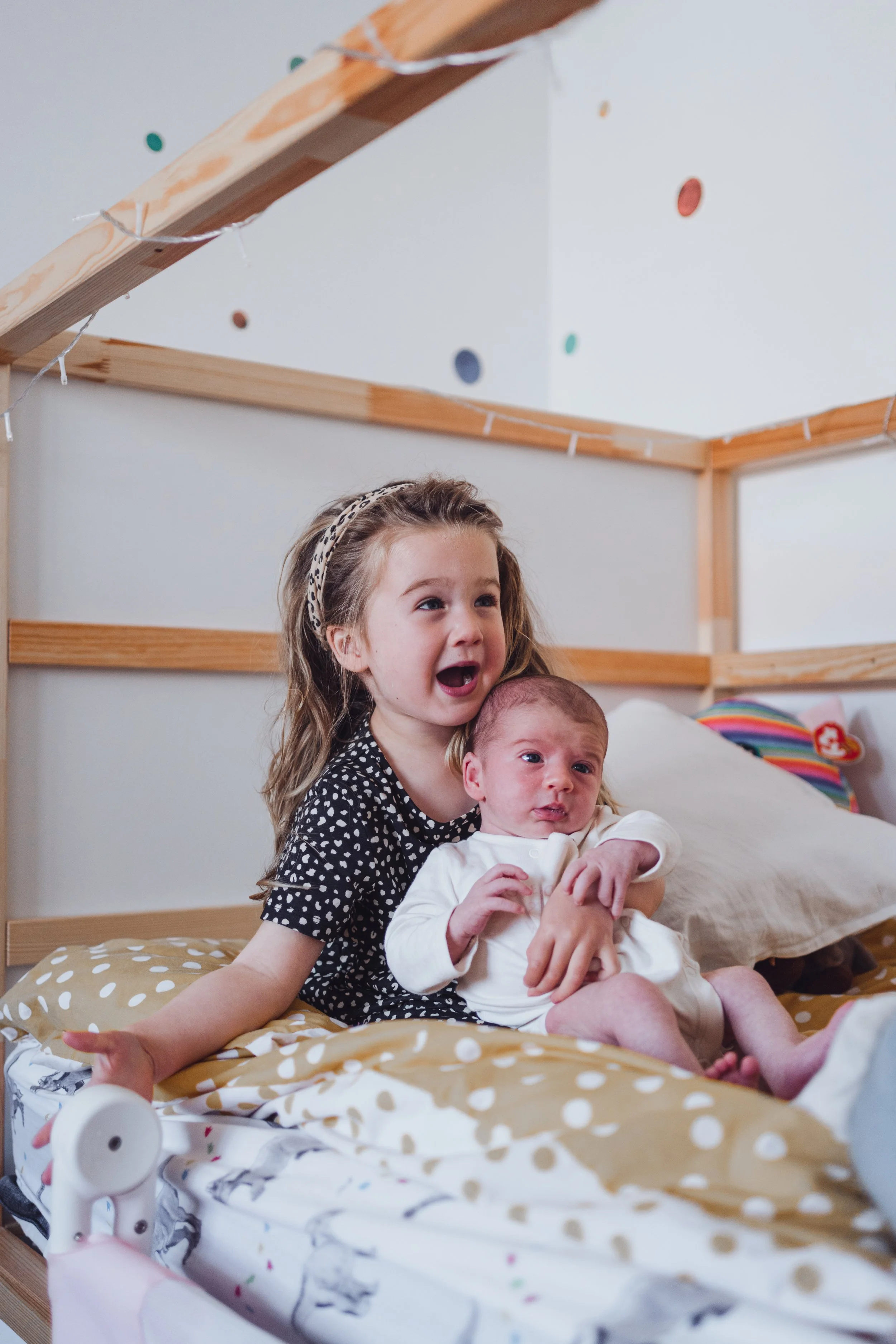 A young girl holding a newborn baby on a bed, inside a bedroom with wooden accents and colorful wall decor.
