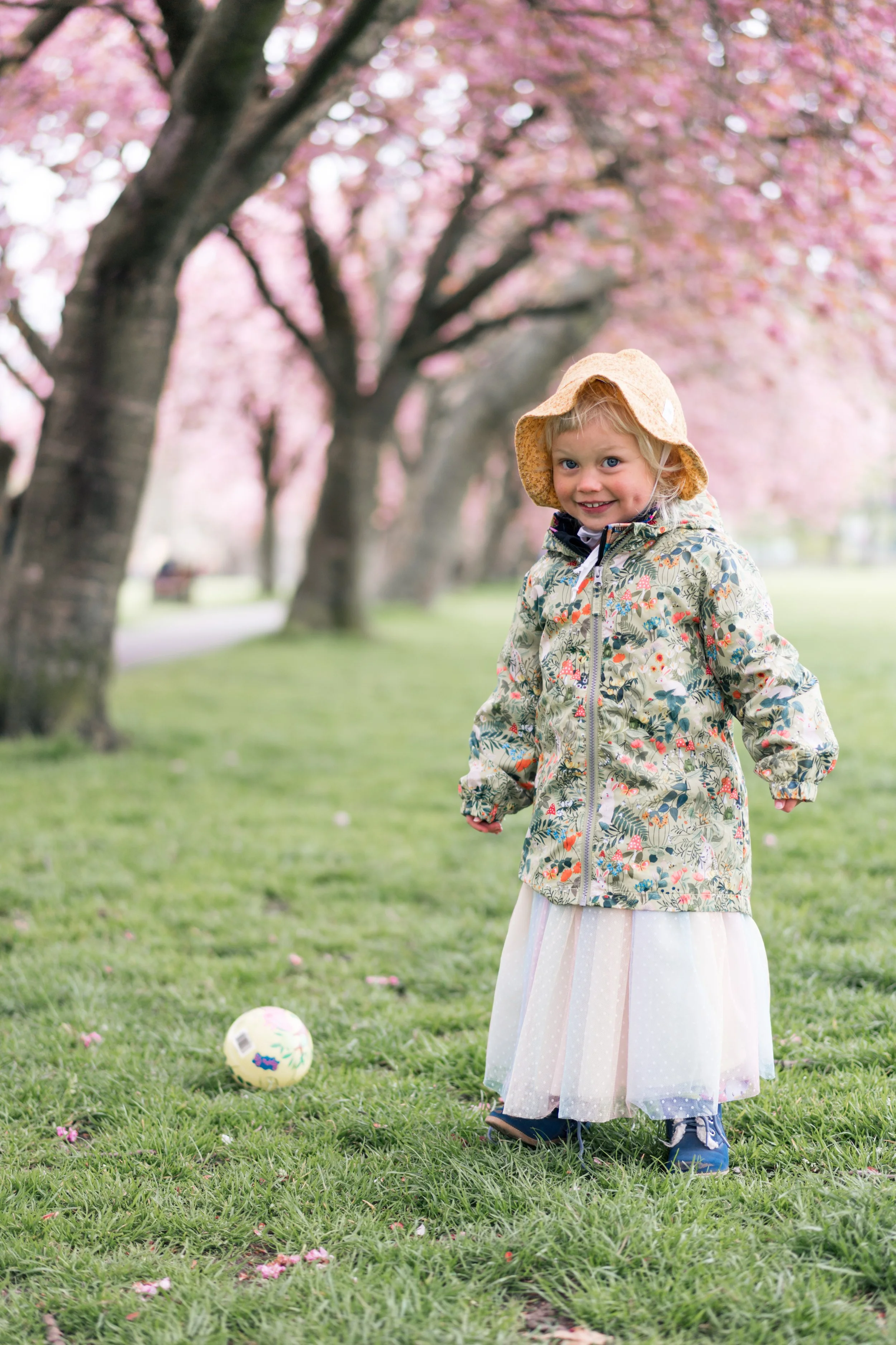 A young girl standing on green grass under pink blossom trees, smiling, wearing a floral jacket, a tulle skirt, a hat, and blue shoes, with a soccer ball nearby.