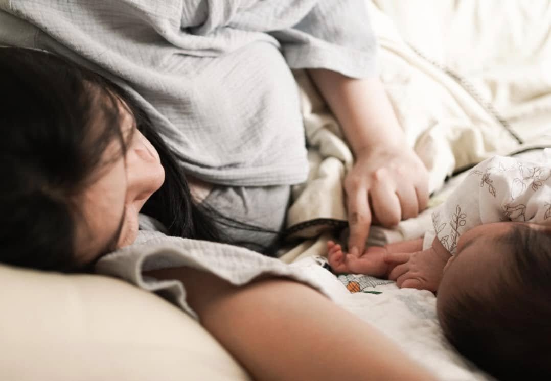 A woman and a baby lying on a bed, holding hands, with the woman smiling and the baby reaching out.