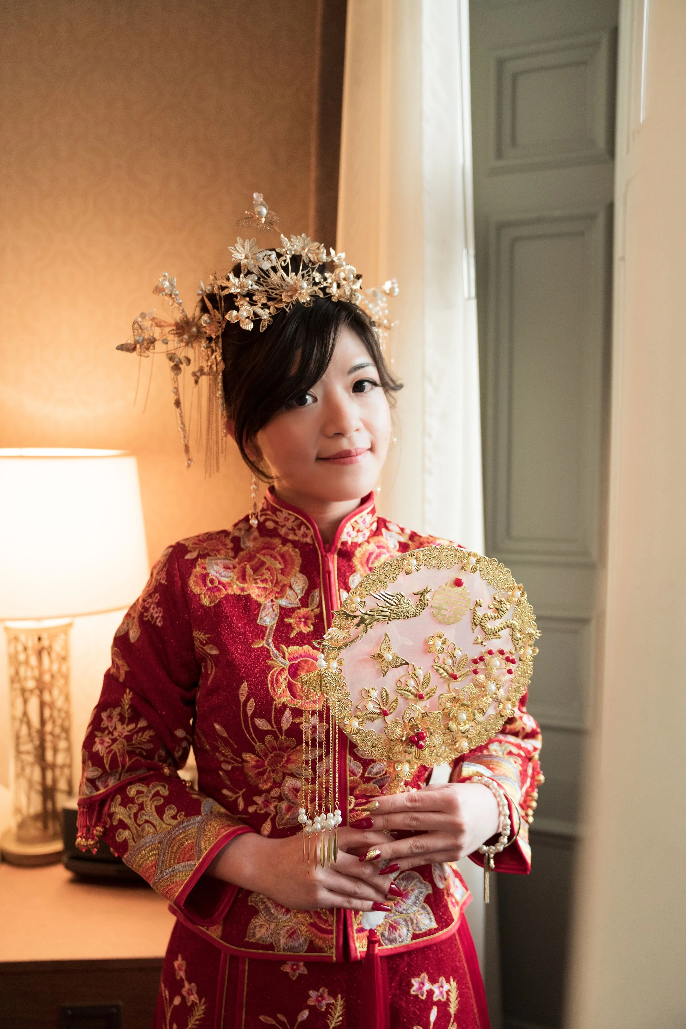 A woman dressed in traditional Chinese wedding attire holding a decorative fan, wearing an ornate floral crown, standing indoors near a lamp and window.