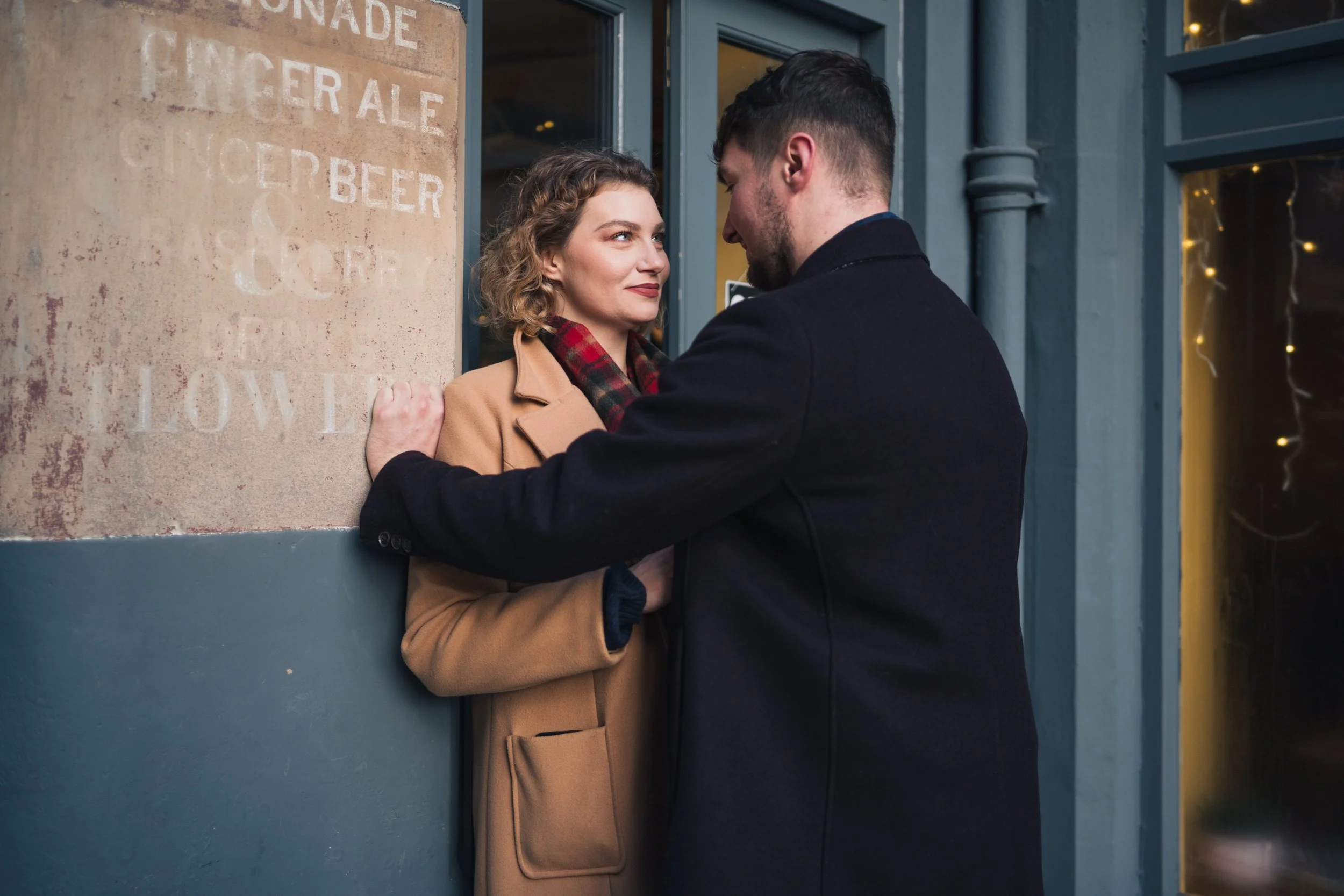 A man and woman are standing close together outside a building, facing each other. The woman is wearing a tan coat with a plaid scarf, and the man is dressed in a dark coat. They are looking into each other's eyes, with the man gently holding the wom
