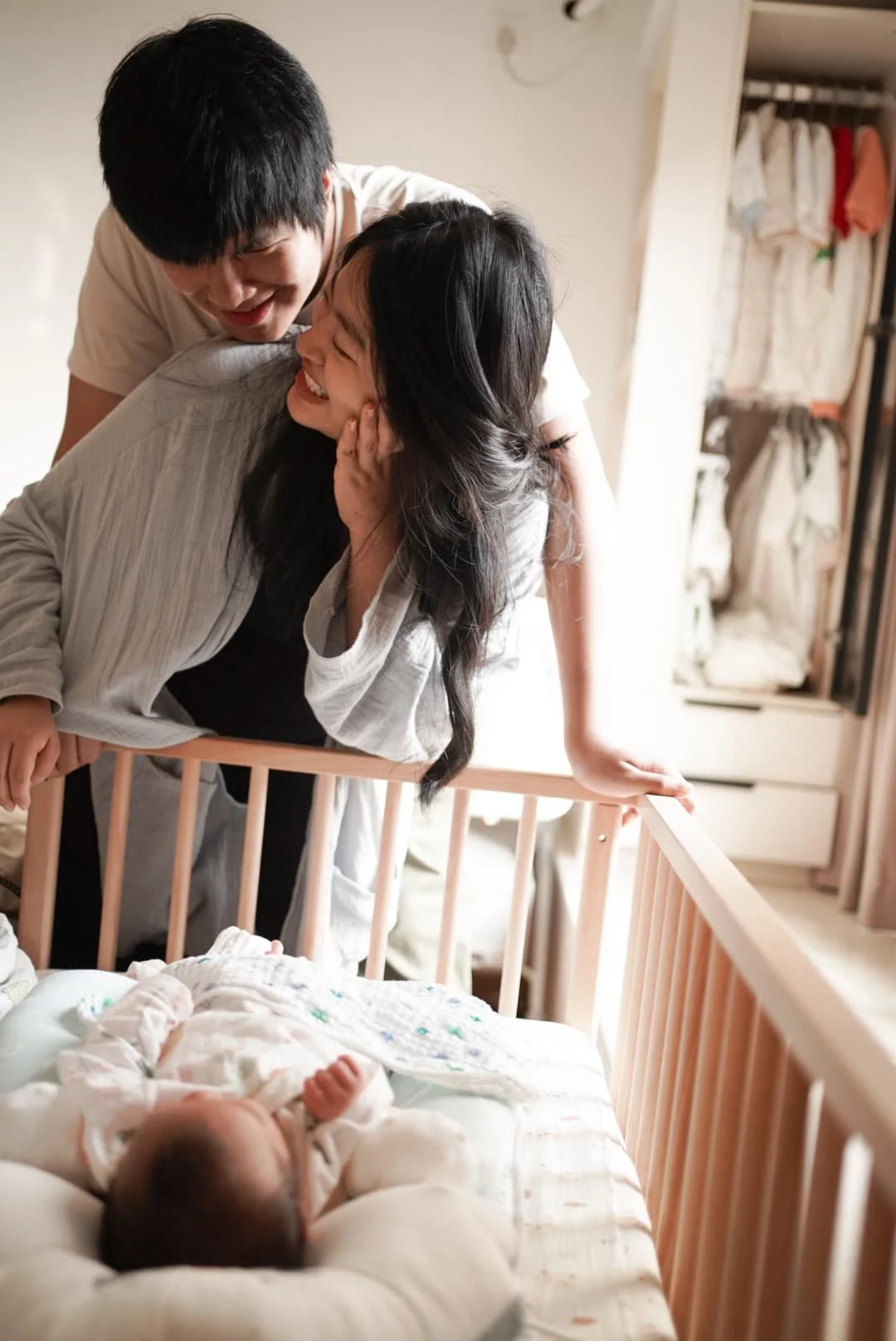 A couple leans over a crib, smiling and laughing at a newborn baby because of the mother and father in the crib.