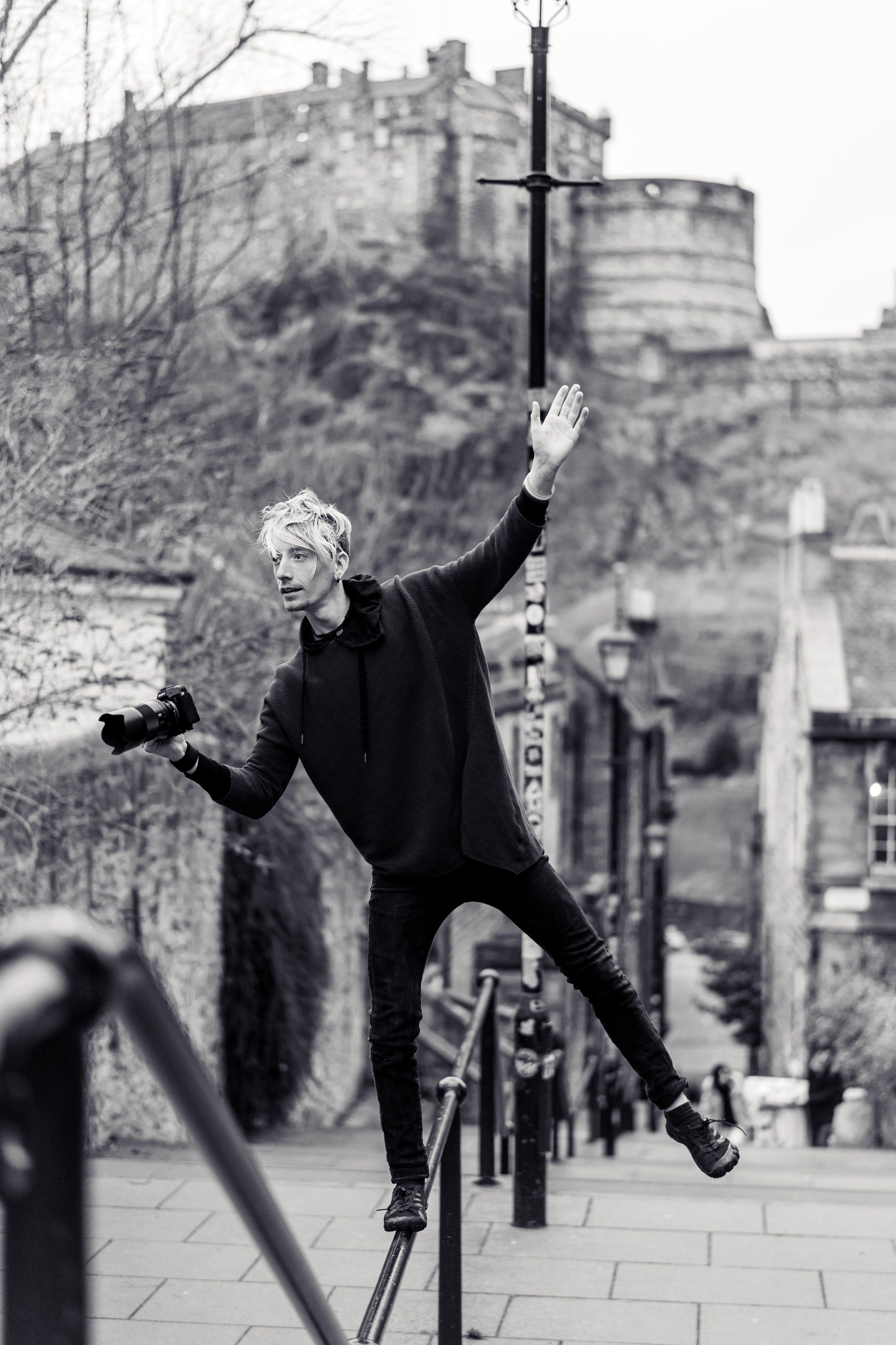 A young man with blonde hair, wearing a dark hoodie, balancing on a railing with arms outstretched, holding a camera in his left hand, in an outdoor urban setting with buildings and a hill in the background.
