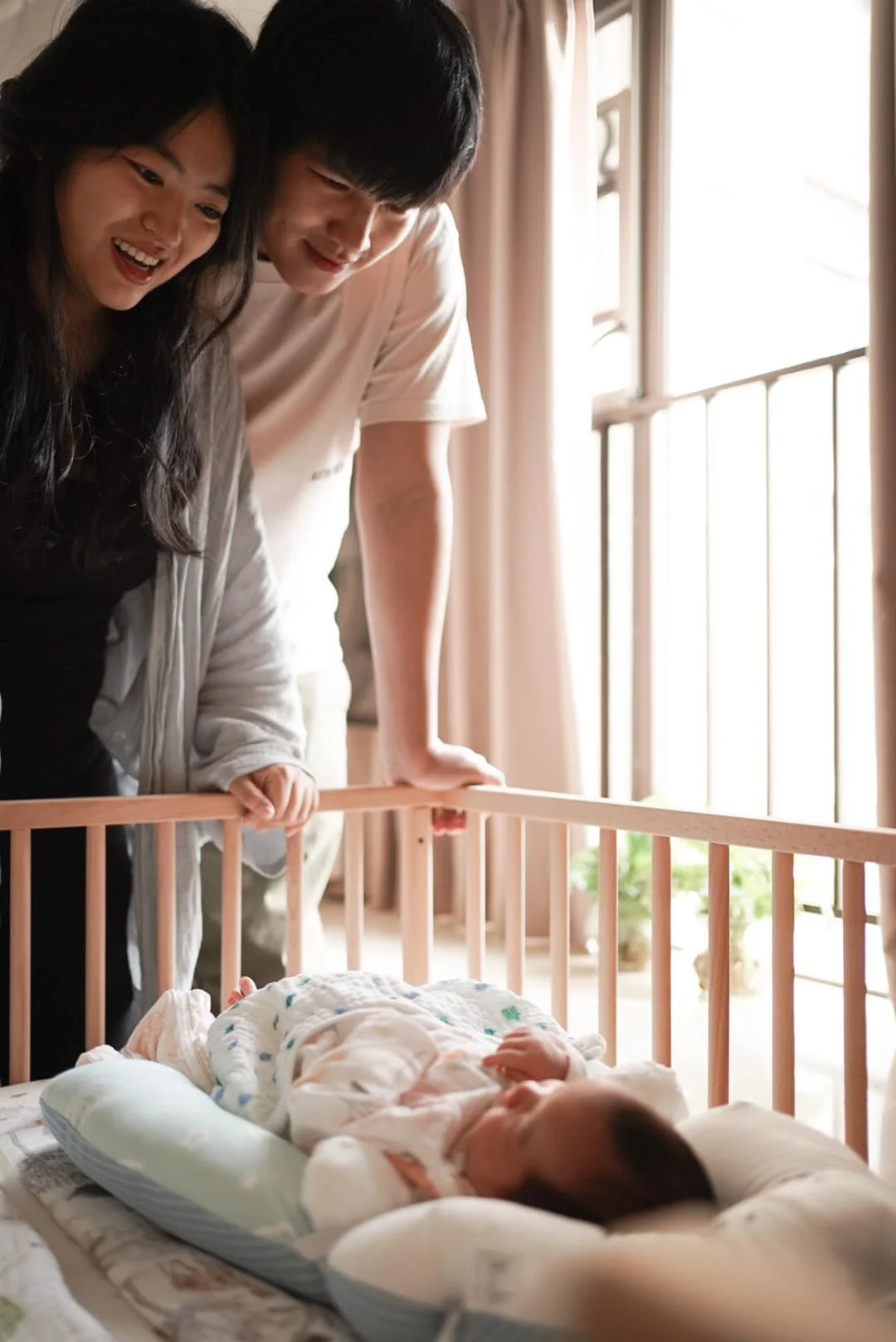 Two adults, a woman and a man, smiling and leaning over a crib to look at a small baby lying inside, with sunlight coming through a window in the background.