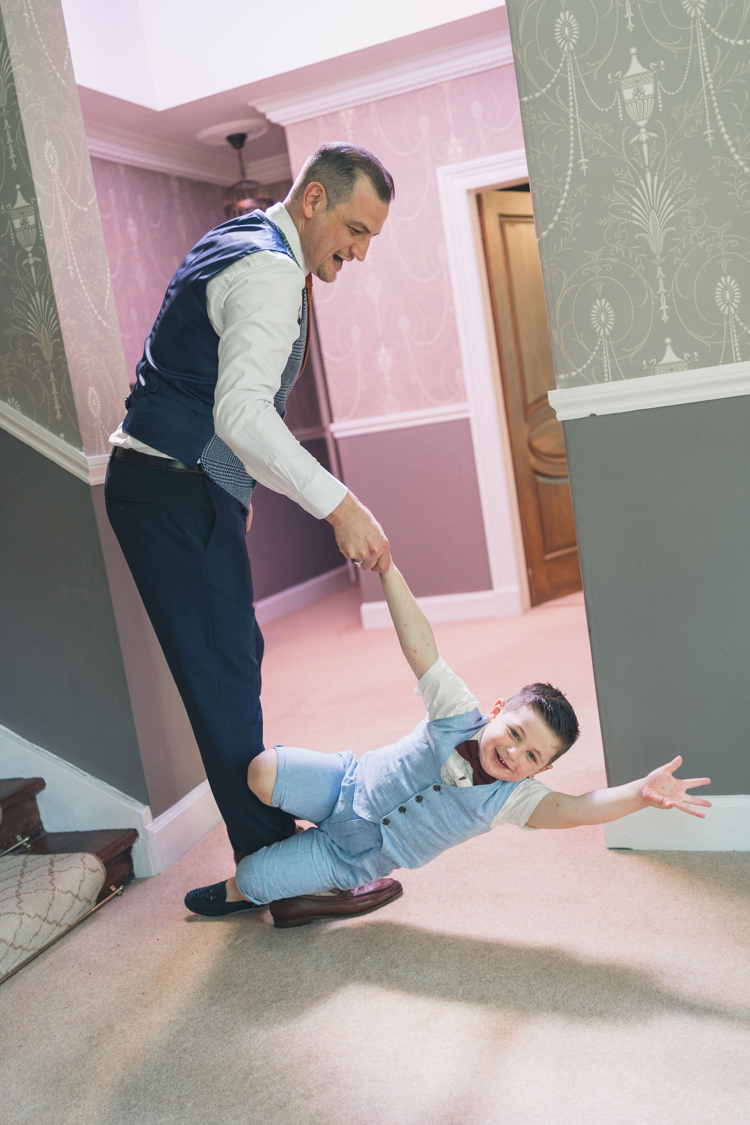 A man and a young boy playing pretend, with the man holding the boy's hand and the boy stretching out on the ground, smiling, inside a house with pink and gray wallpaper and a staircase.