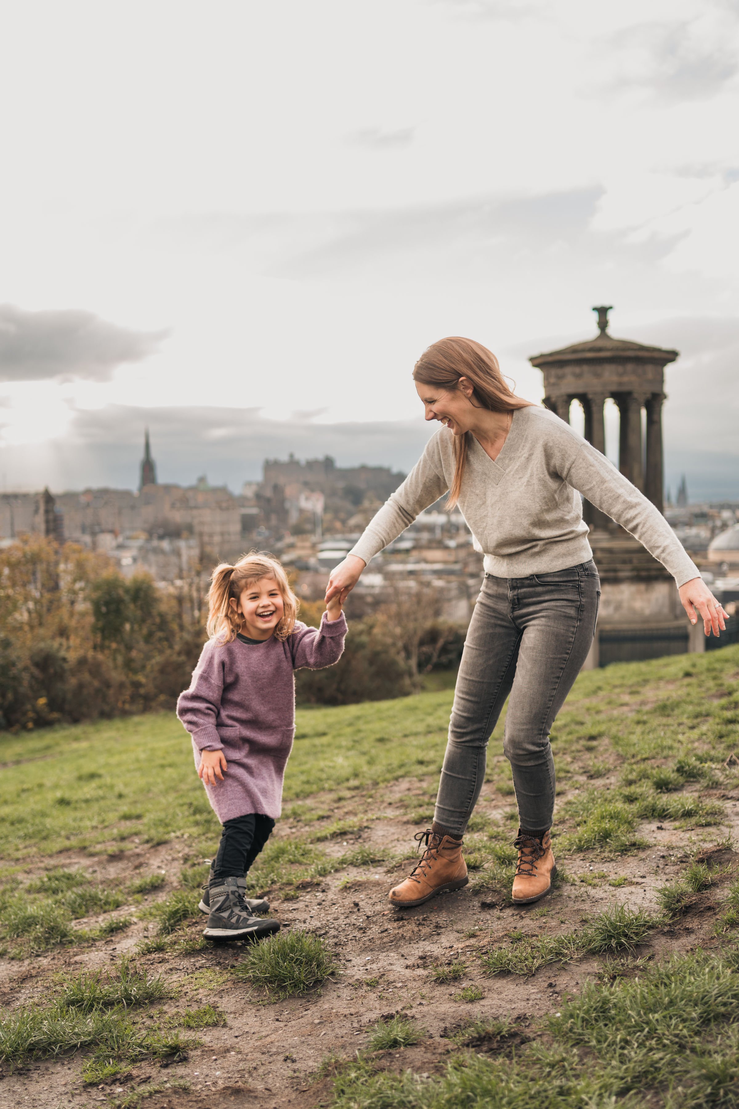 A woman and a young girl, likely mother and daughter, are playing and dancing on a grassy hill with a cityscape and historic structures in the background. The woman is holding the girl's hand, and both are smiling and laughing.