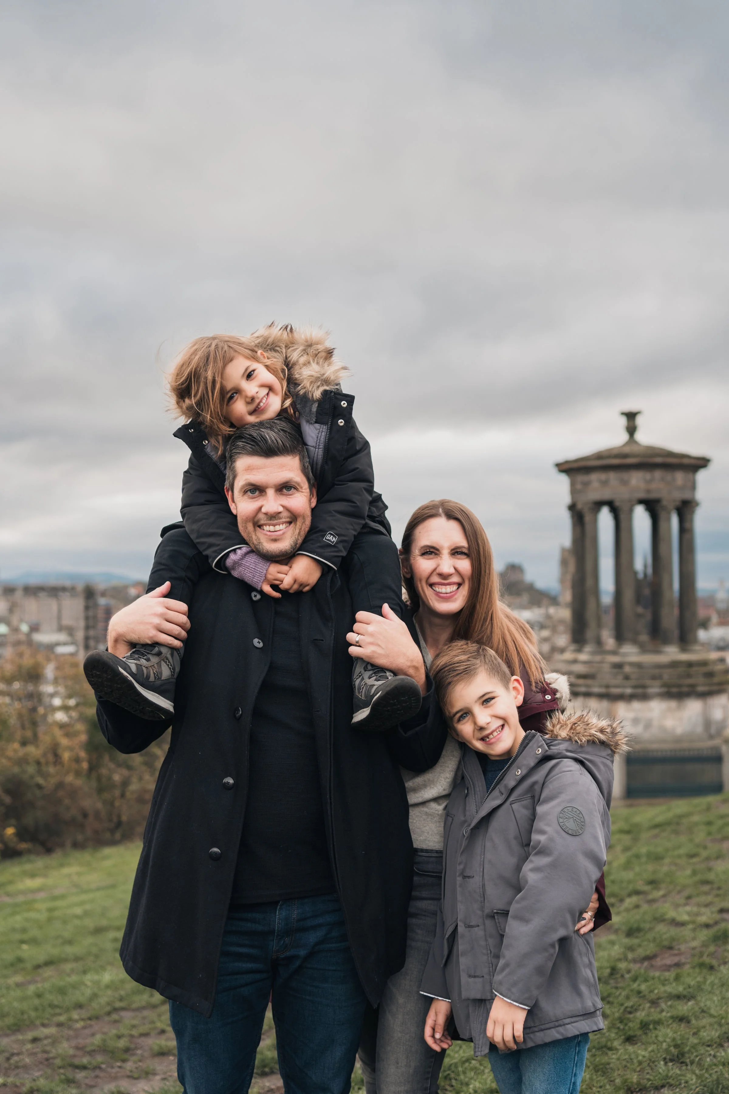 A happy family of four posing outdoors with a historical monument in the background, under cloudy sky.