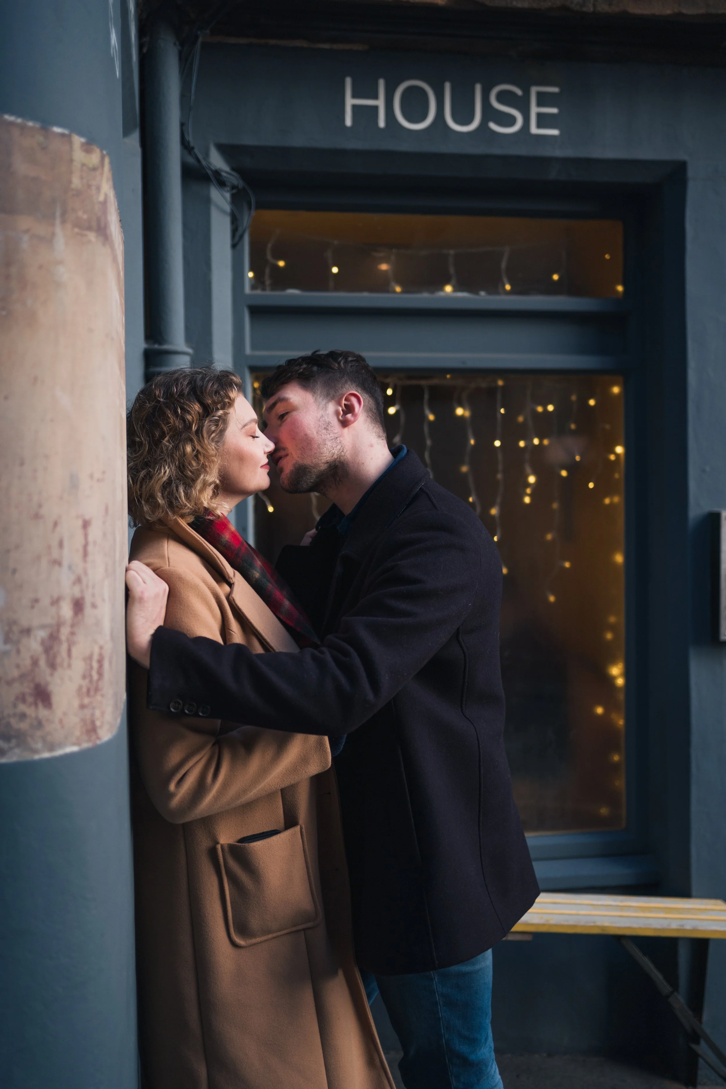A man and woman share an intimate moment outside a building labeled "HOUSE," with festive string lights illuminating the background.