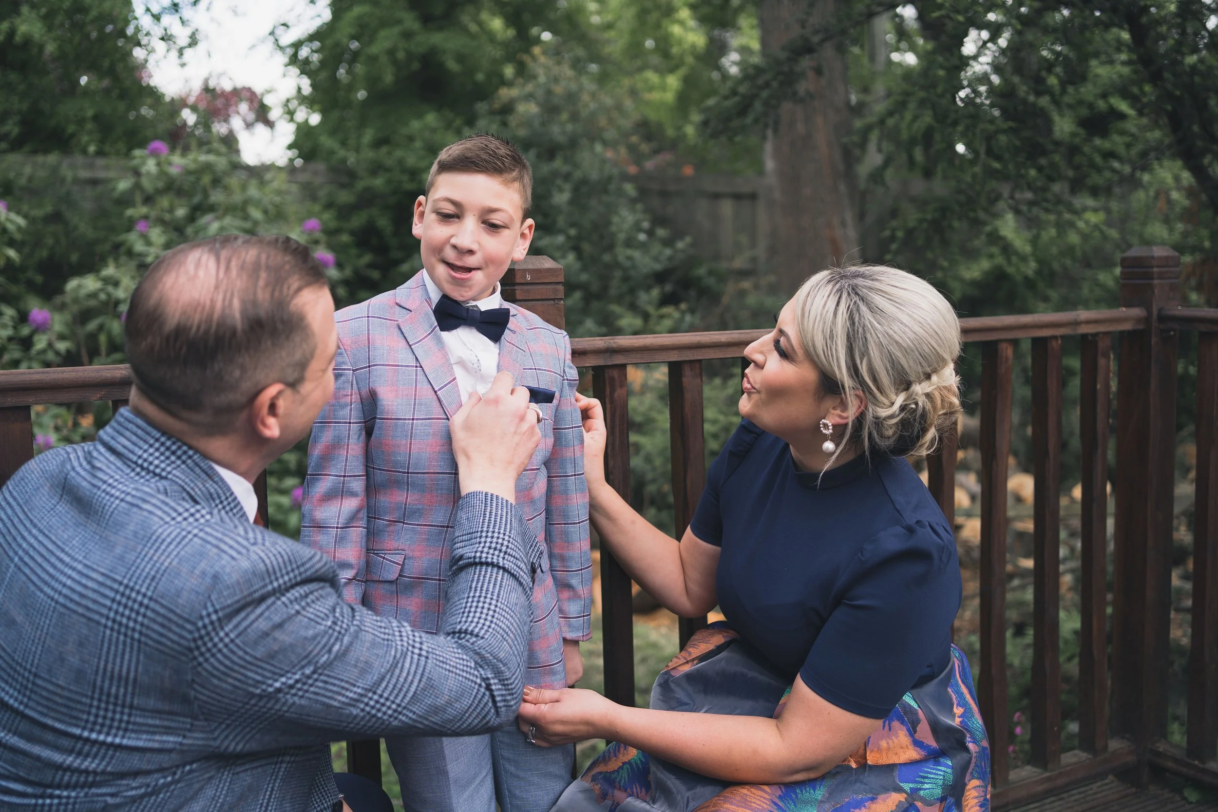 A boy dressed in a plaid suit and bow tie is being helped with his outfit by a man and a woman, likely parents, on a wooden deck outdoors with trees and foliage in the background.