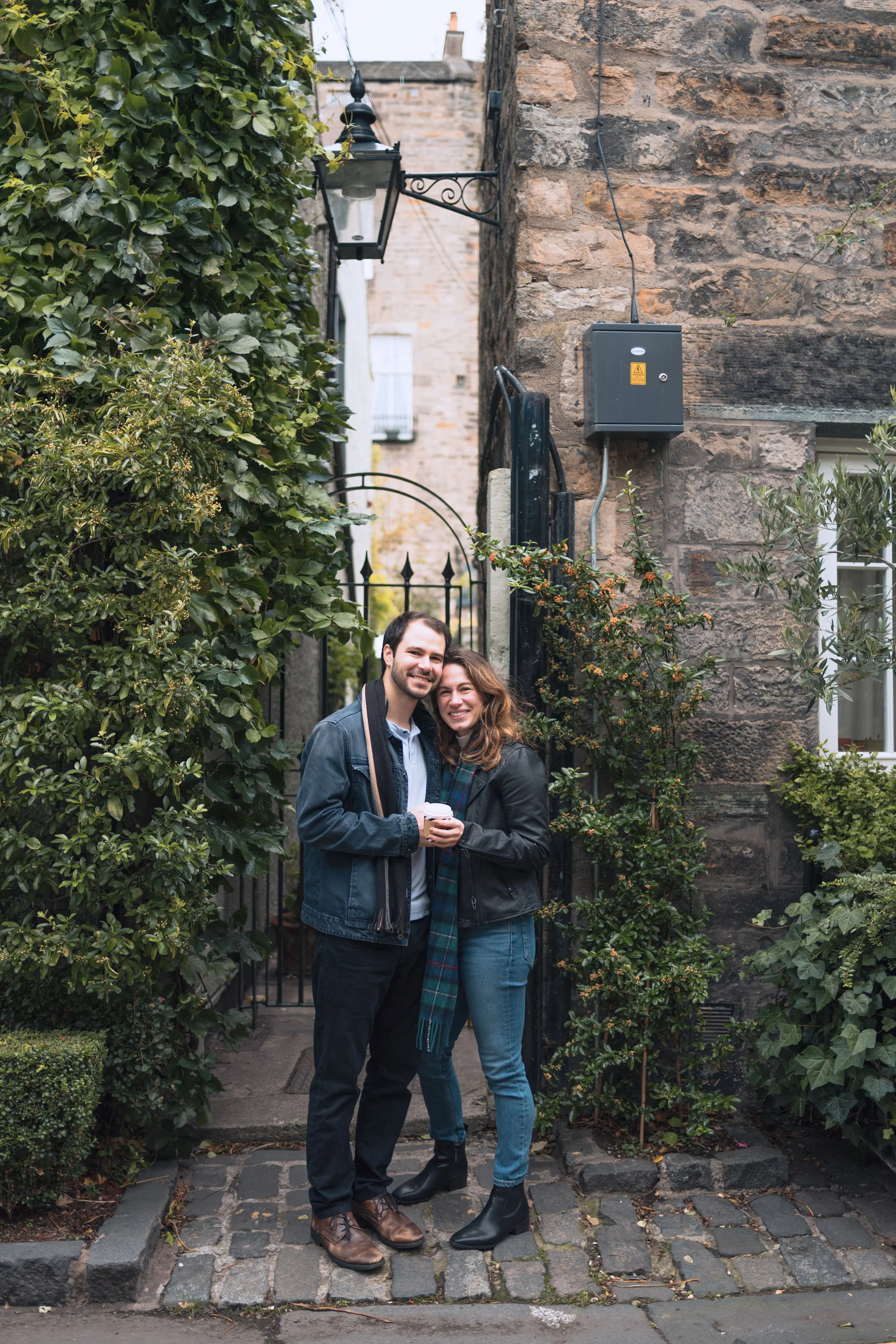 A smiling couple standing close together on a cobblestone street, holding a cup of coffee, surrounded by green bushes and old brick buildings.