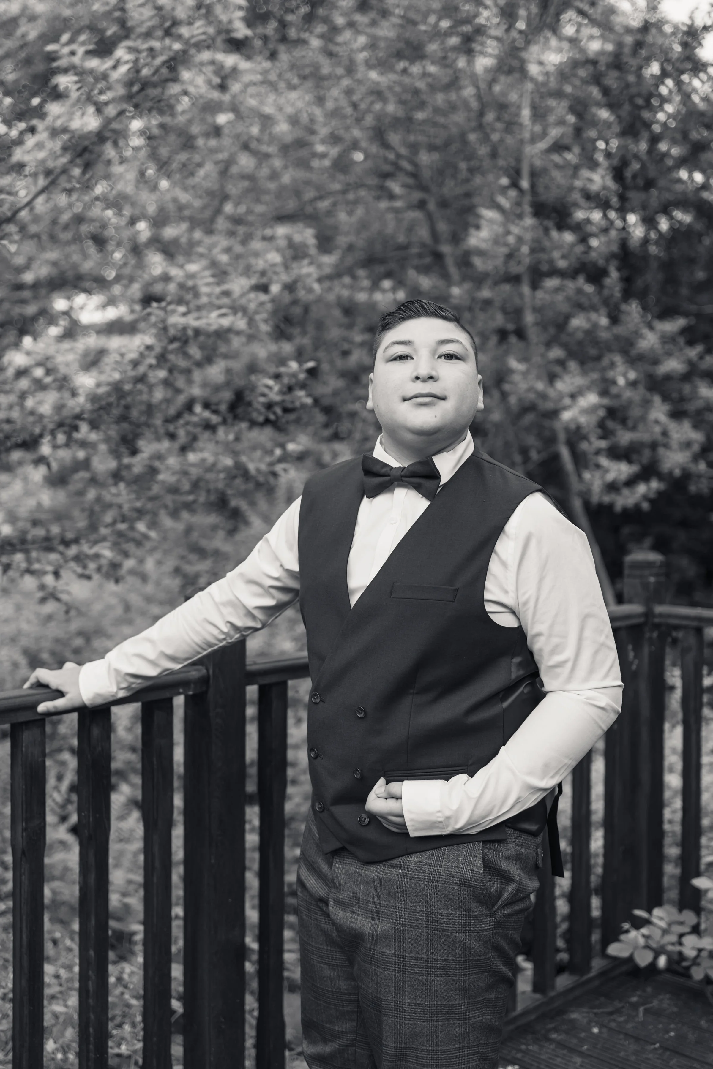 A young man wearing formal attire including a tuxedo vest, bow tie, and dress shirt, standing outdoors on a wooden bridge with a railing, with trees and foliage in the background, in black and white.
