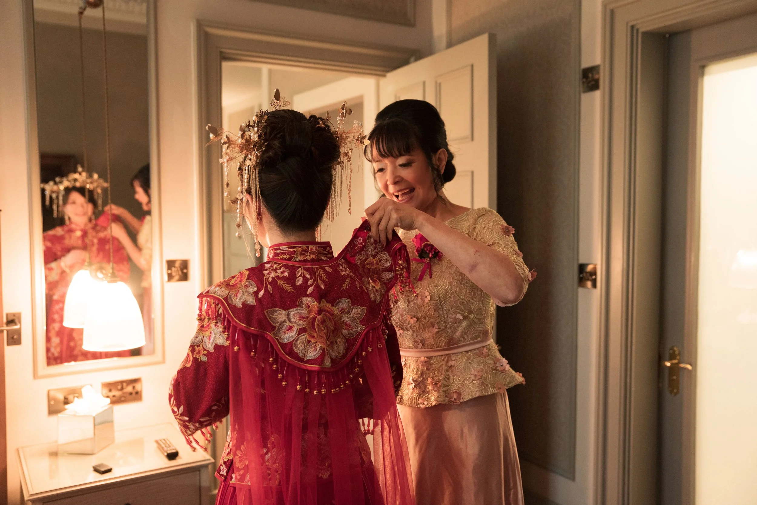 A woman helping another woman into a traditional Chinese wedding dress in a warmly lit room. The woman being helped is dressed in a red embroidered gown, and the woman assisting is wearing a gold dress.