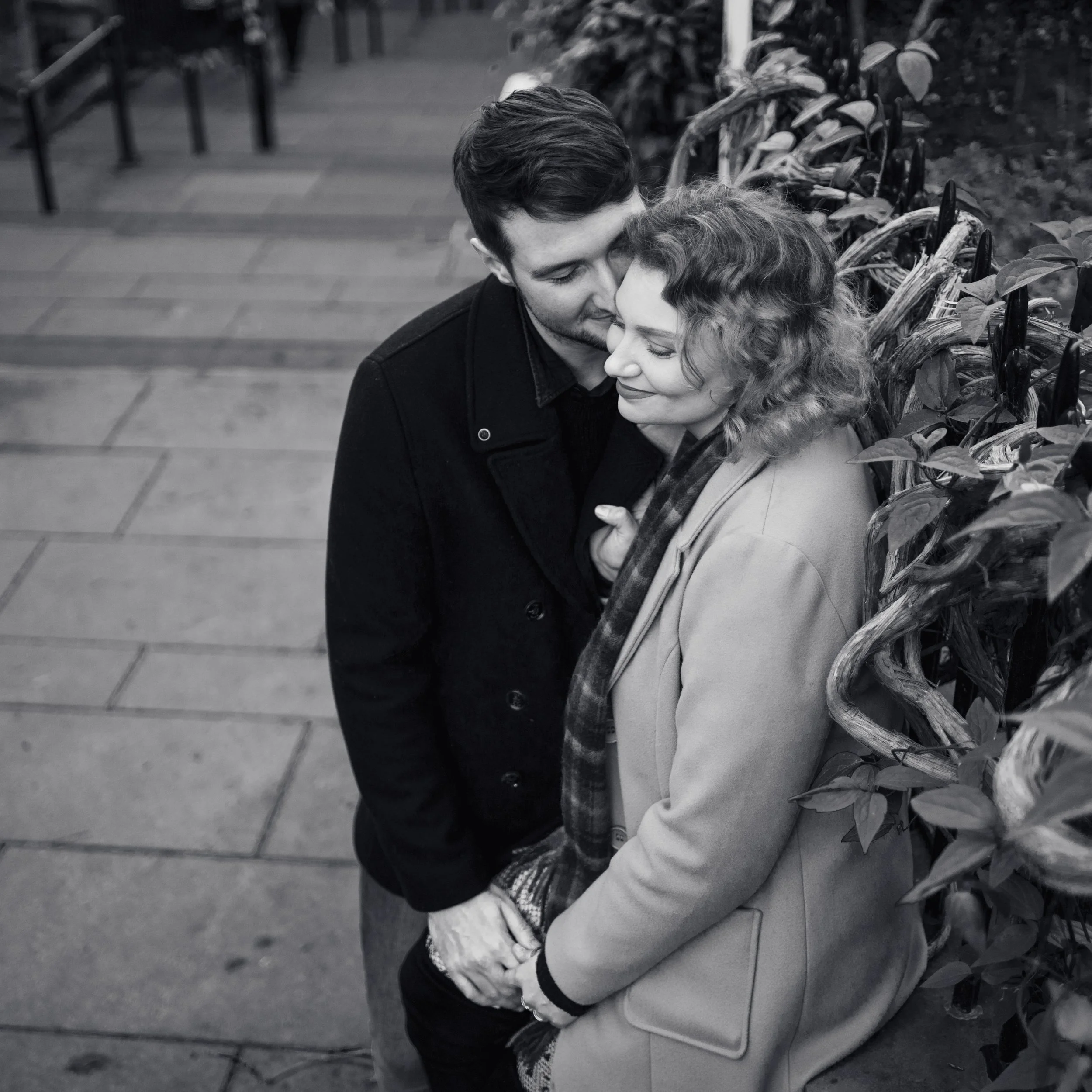 A black and white photo of a couple standing close together on a sidewalk, with the woman leaning against a wooden fence and the man whispering to her affectionately.