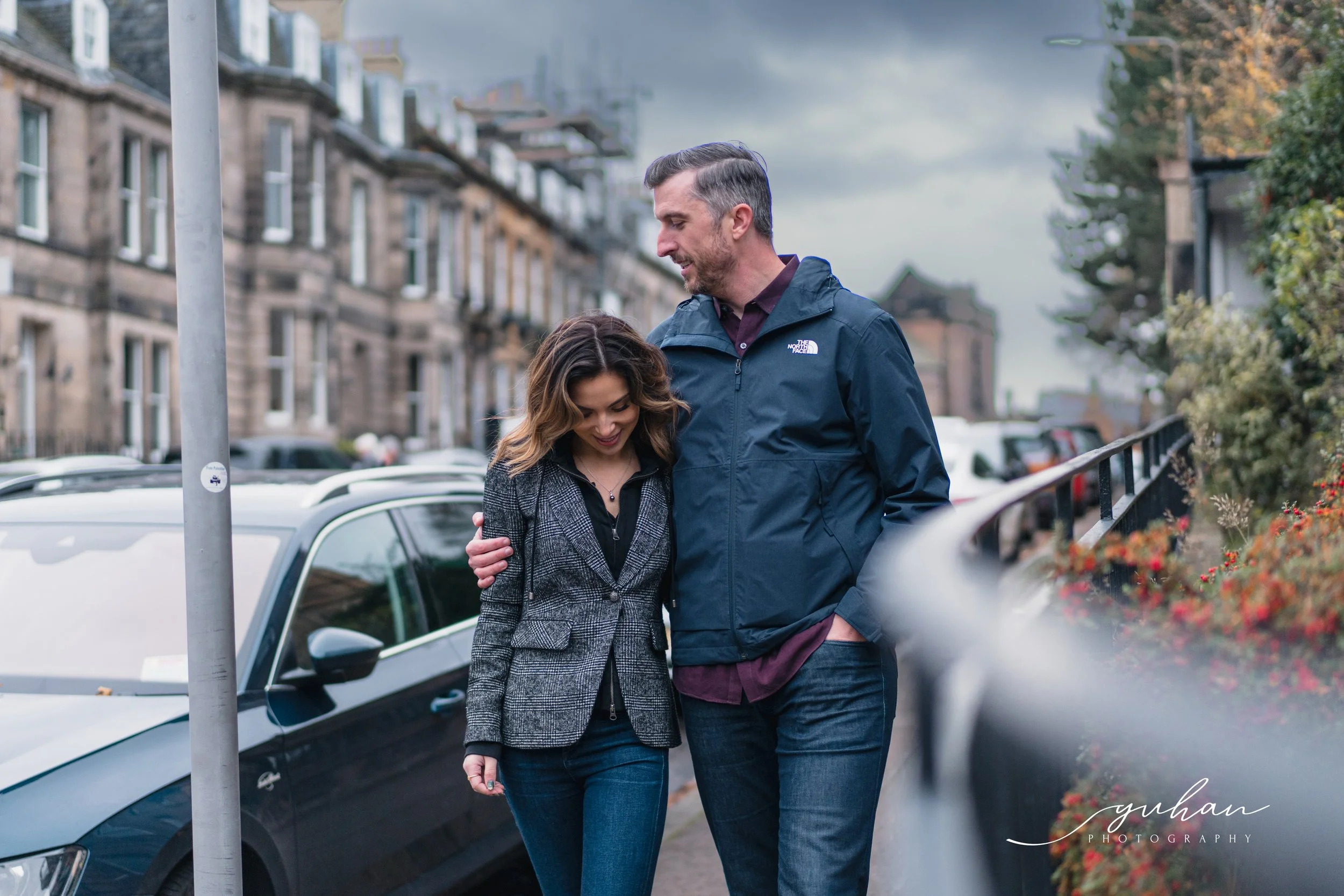 A man and a woman walking together outdoors on a city street, smiling and looking down, with the man’s arm around the woman’s shoulder, before a background of parked cars and residential buildings.
