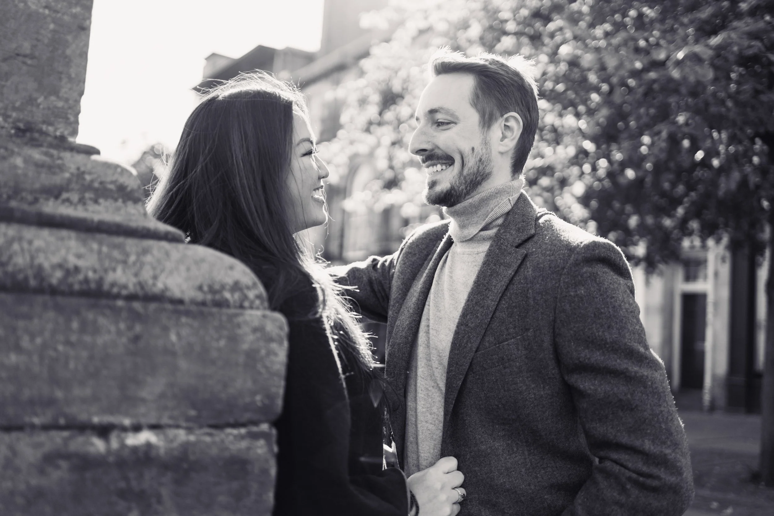 A black and white photograph of a smiling man and woman looking at each other outdoors, with the woman leaning against a brick wall and trees in the background.