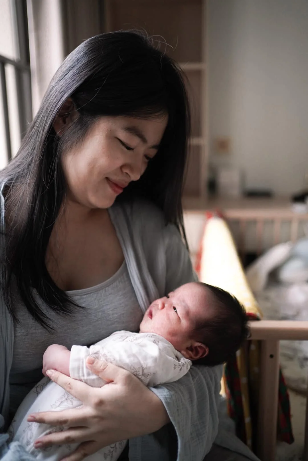 A woman holding a newborn baby in a cozy indoor setting near a window with curtains